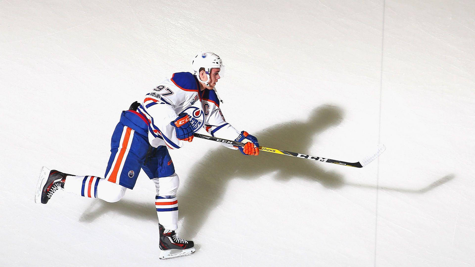 hockey 桌布 壁紙 — 一名身穿藍、橙、白色球衣的冰上曲棍球運動員在冰場上動態滑行，手持球桿，投下長長的影子。