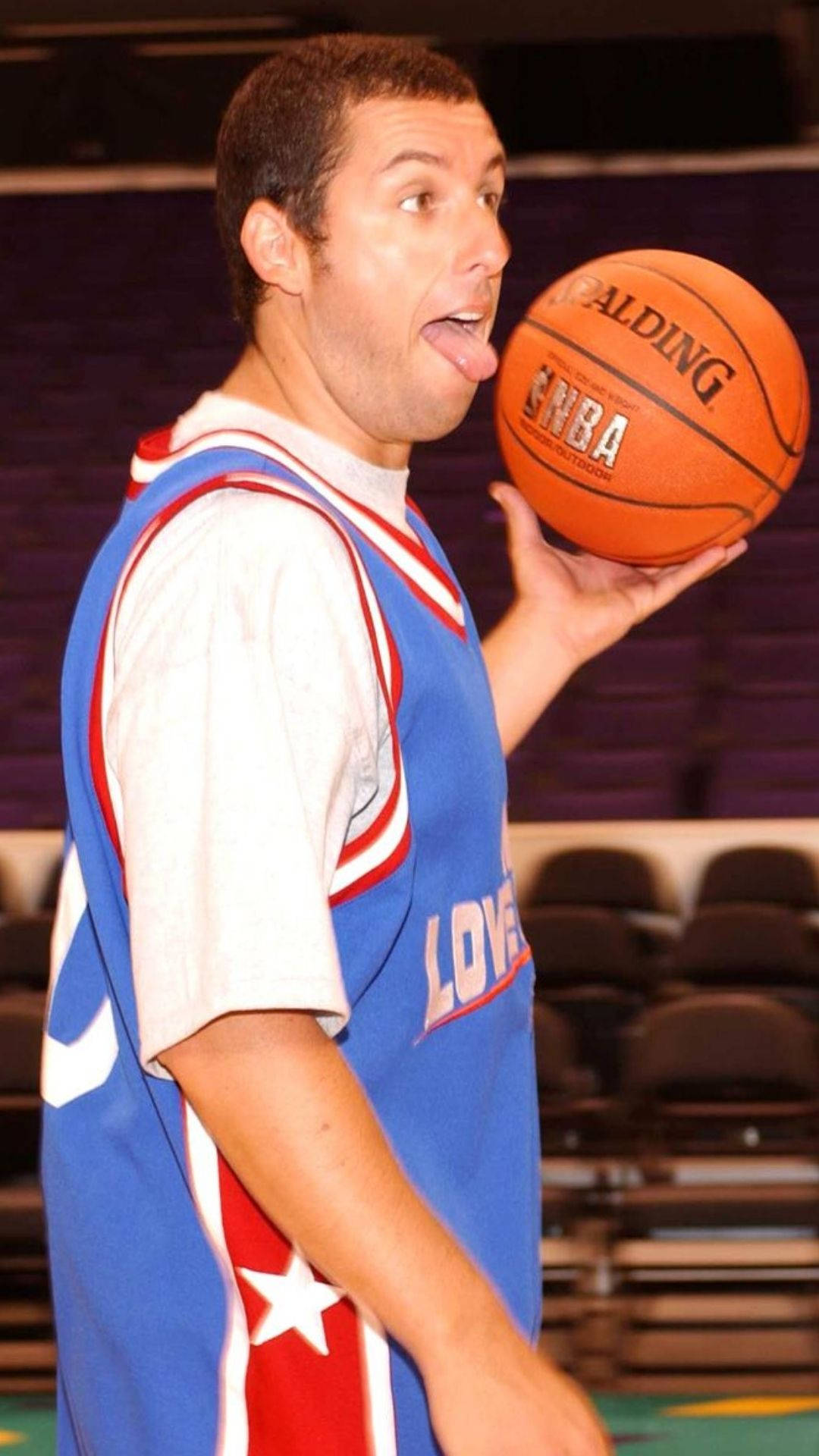 Adam Sandler 桌布 壁紙 — 一名身穿藍、紅、白三色籃球球衣的男子，調皮地伸出舌頭，同時拿著一個橙色的 Spalding NBA 籃球。