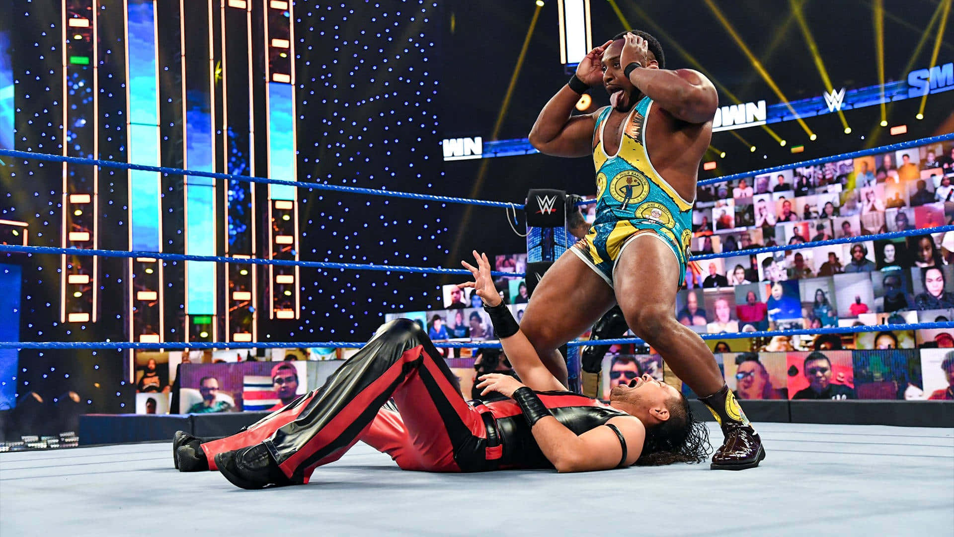 WWE 桌布 壁紙 — 一名身穿藍、黃、金色服裝的摔角手，在燈光明亮的摔角場內，將身穿紅黑皮褲的對手壓制住。