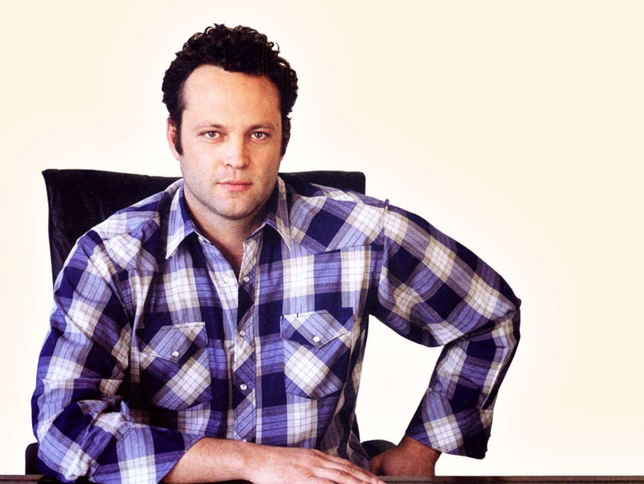 Vince Vaughn 桌布 壁紙 — 一名身穿藍白格紋襯衫的男子自信地坐在辦公桌前，背景是純色米白色。