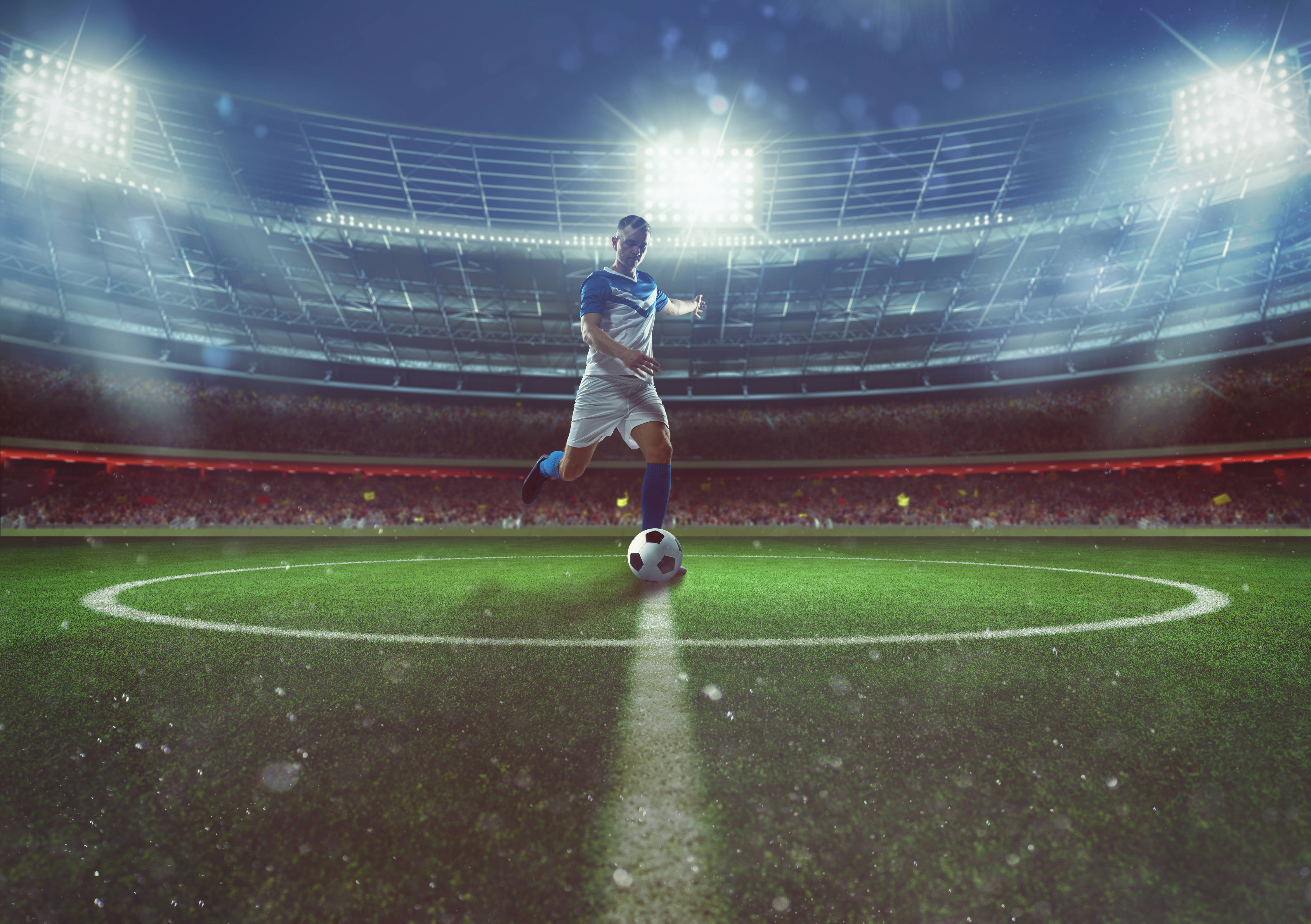 Football Field 桌布 壁紙 — 一名身穿藍白球衣的足球運動員在明亮的球場燈光下，於充滿活力的綠色草地上運球，背景是座無虛席的人群。