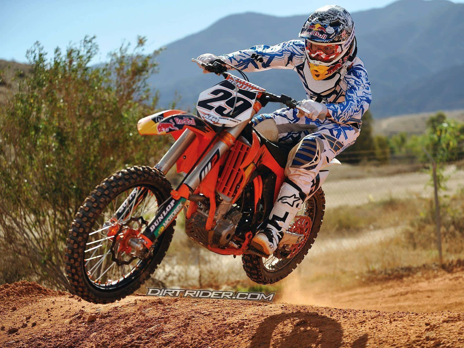 Ktm Bike 桌布 壁紙 — 一名身穿藍白色裝備的越野摩托車手，騎著橙色越野摩托車，在塵土飛揚的土坡上空飛躍，背景是山脈。