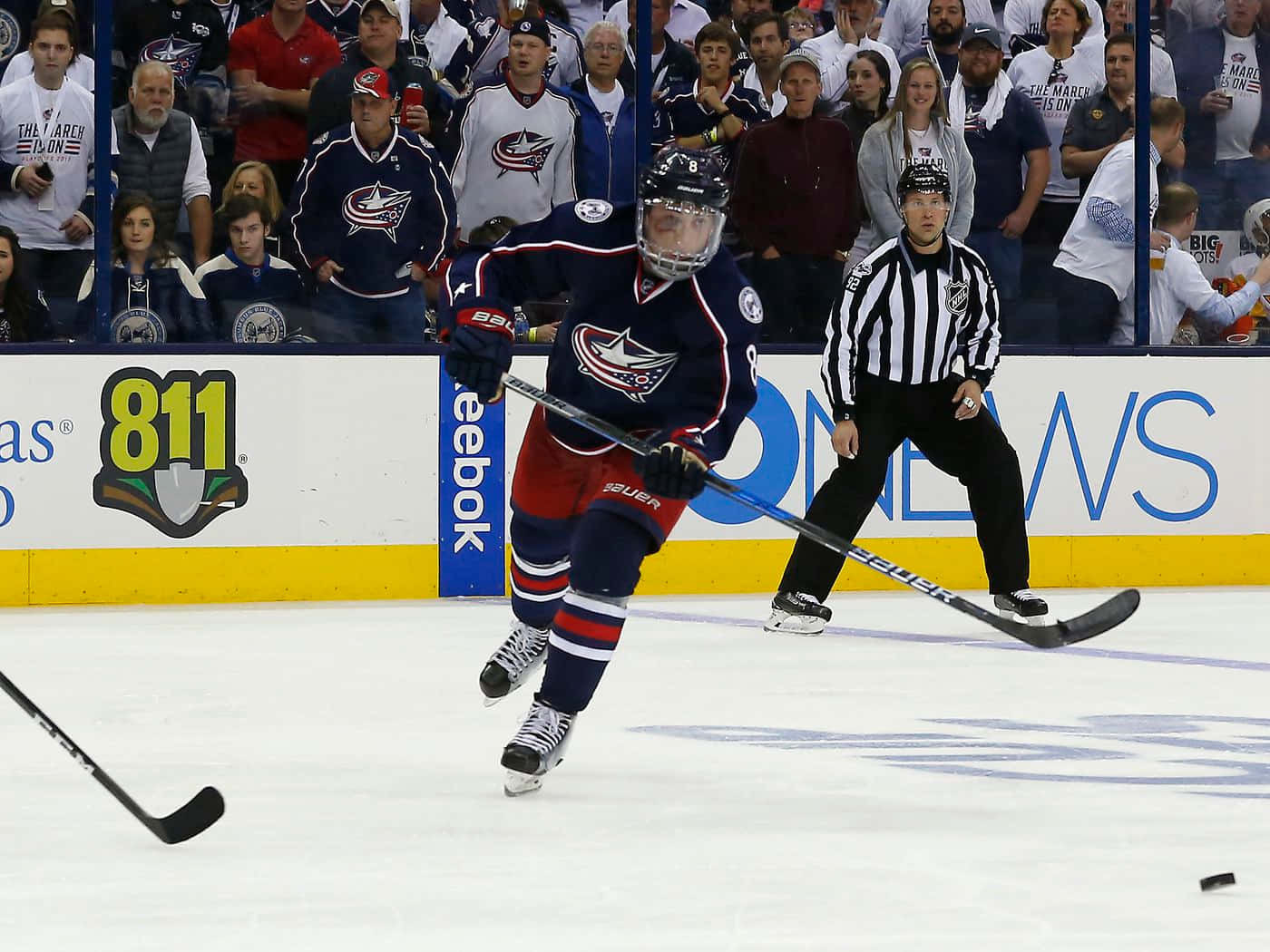 運動 桌布 壁紙 — 一名身穿藍紅球衣的冰球運動員帶著冰球滑行，裁判在激烈的比賽中密切注視。