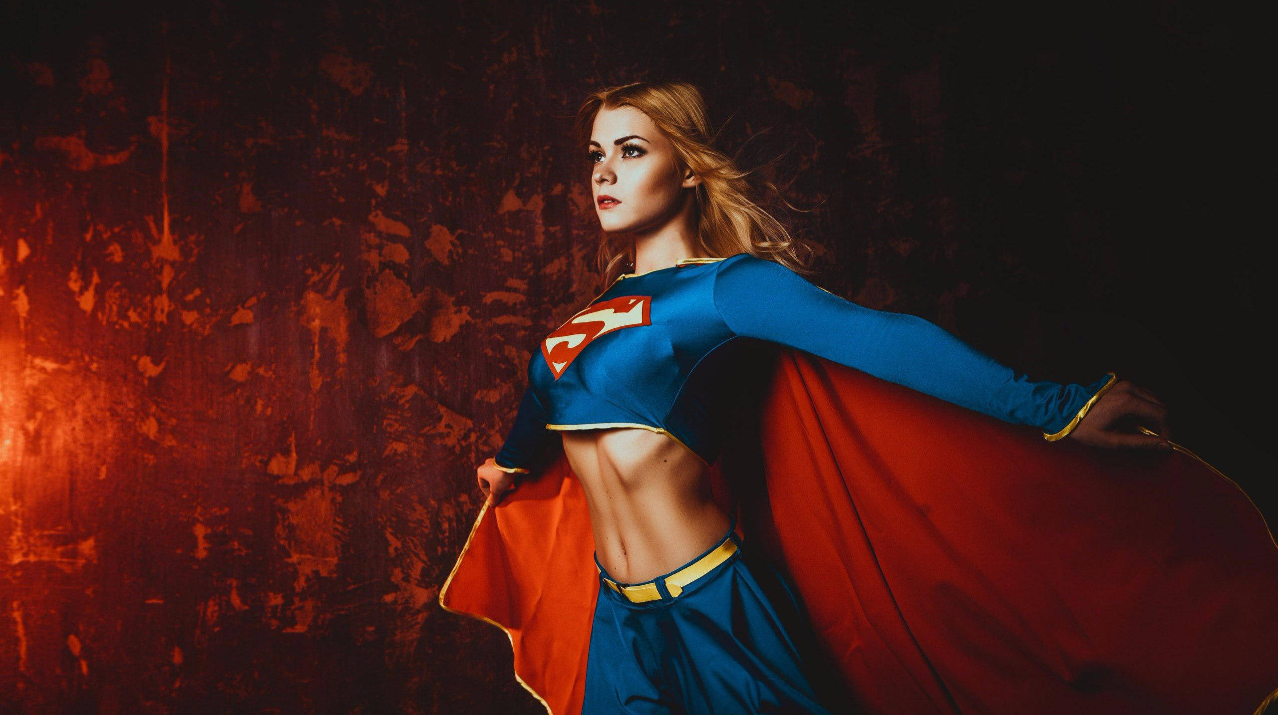 Supergirl 桌布 壁紙 — 一名身穿藍紅配色超級女孩服裝的女性，自信地站在紋理火紅的牆壁前，搭配戲劇性的燈光。