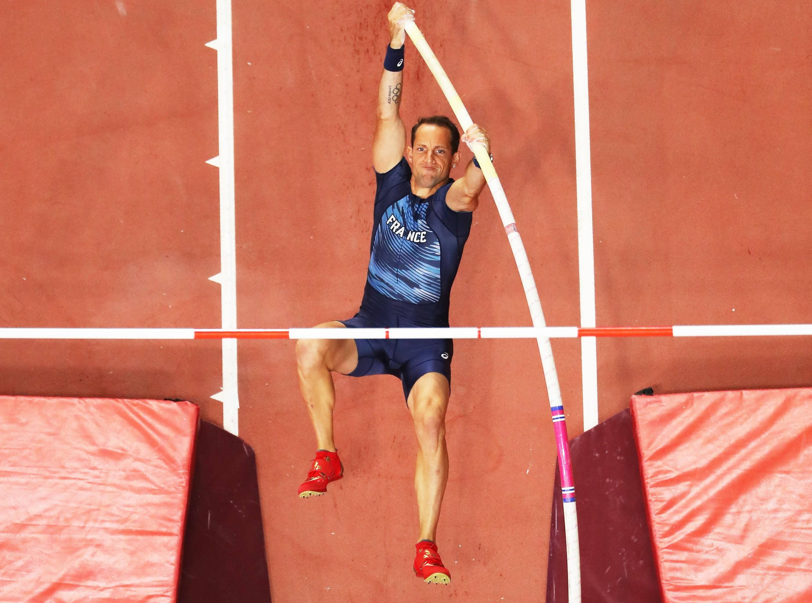Athletic 桌布 壁紙 — 一名身穿藍色「FRANCE」制服的男子撐竿跳選手，在紅色跑道背景下專注地越過橫桿。