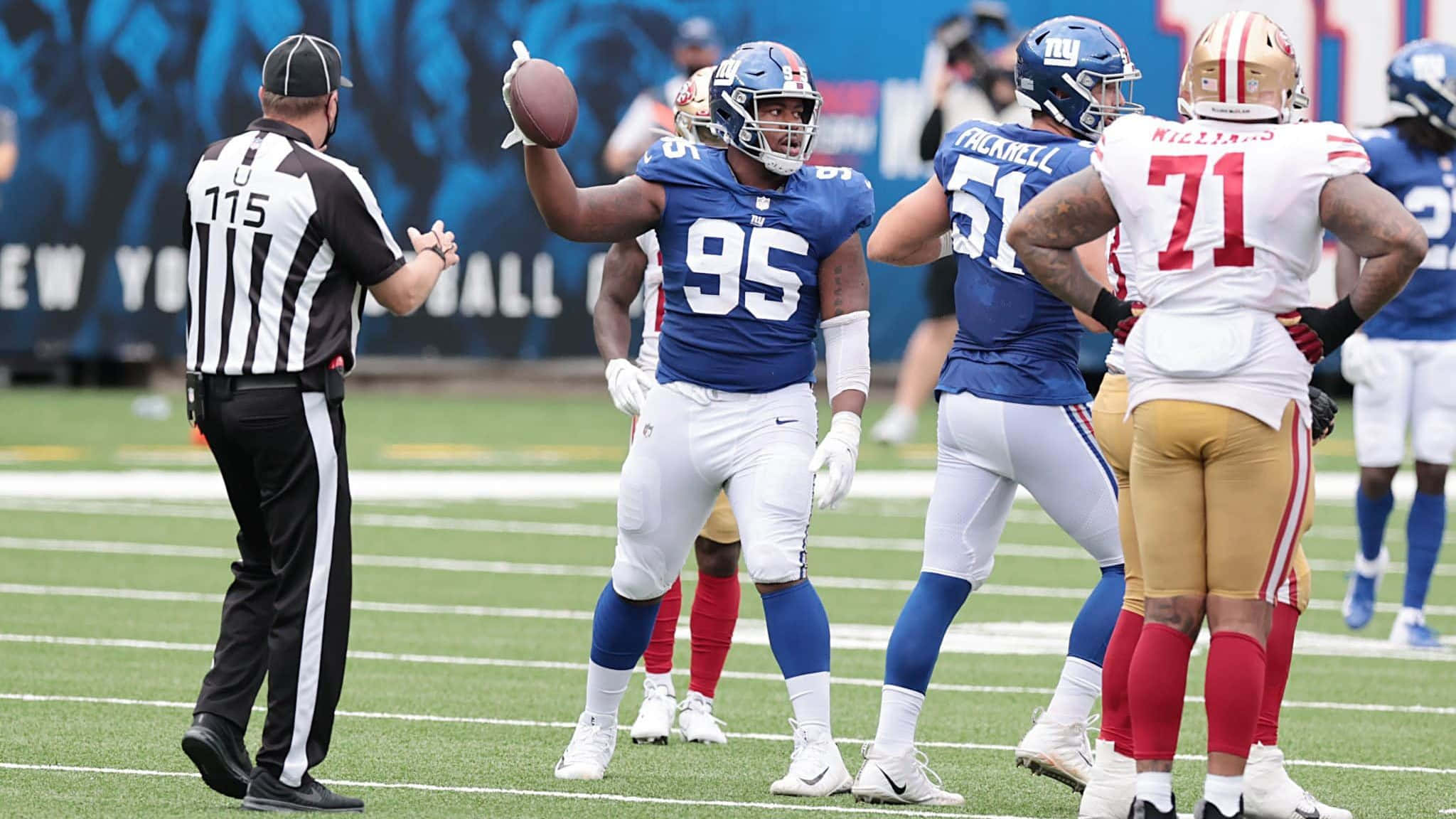 New York Giants 桌布 壁紙 — 一名身穿藍色95號球衣的美式足球員高舉足球，裁判和對方球員站在附近的綠色球場上。