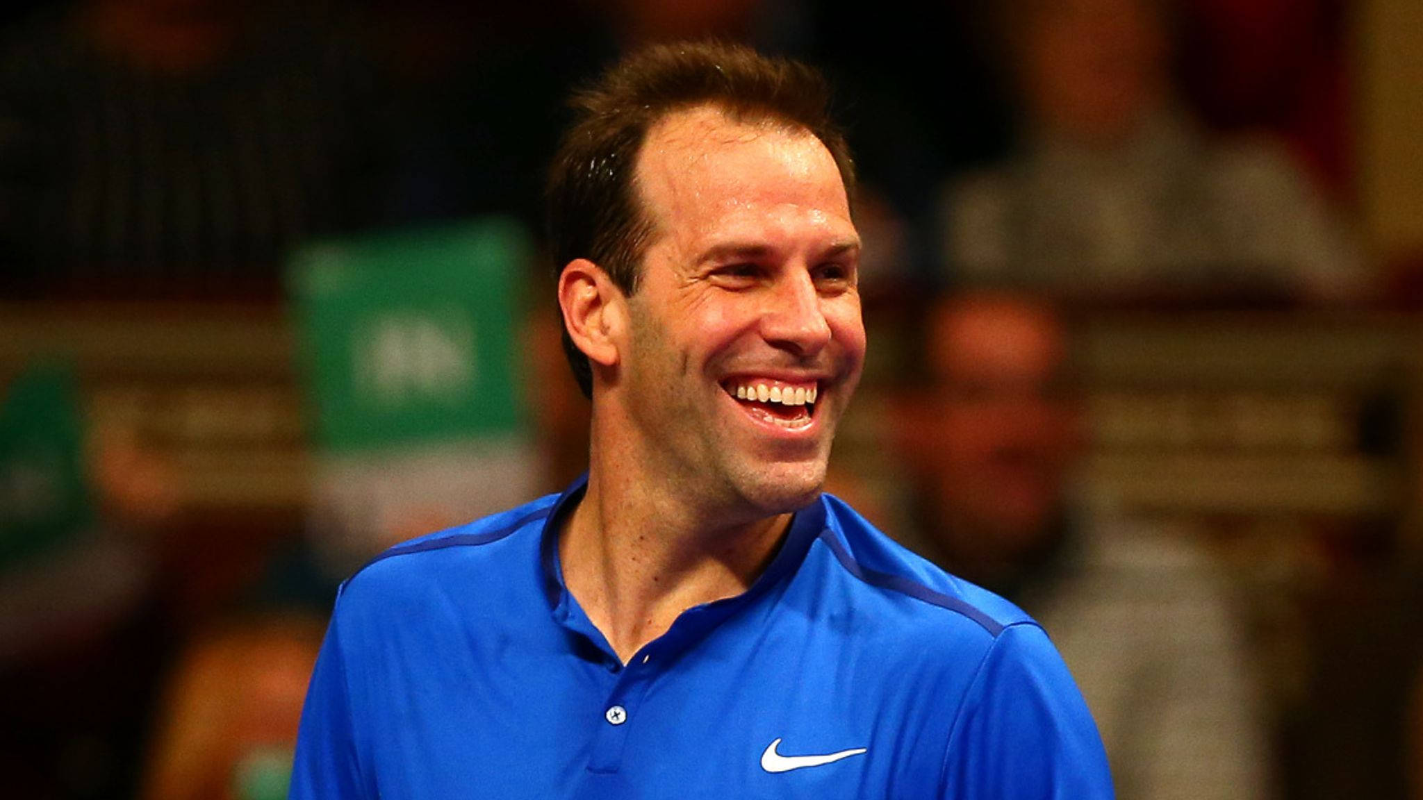 運動 桌布 壁紙 — 一名身穿藍色Nike球衣的網球選手在室內比賽中開懷大笑，背景是模糊的觀眾。