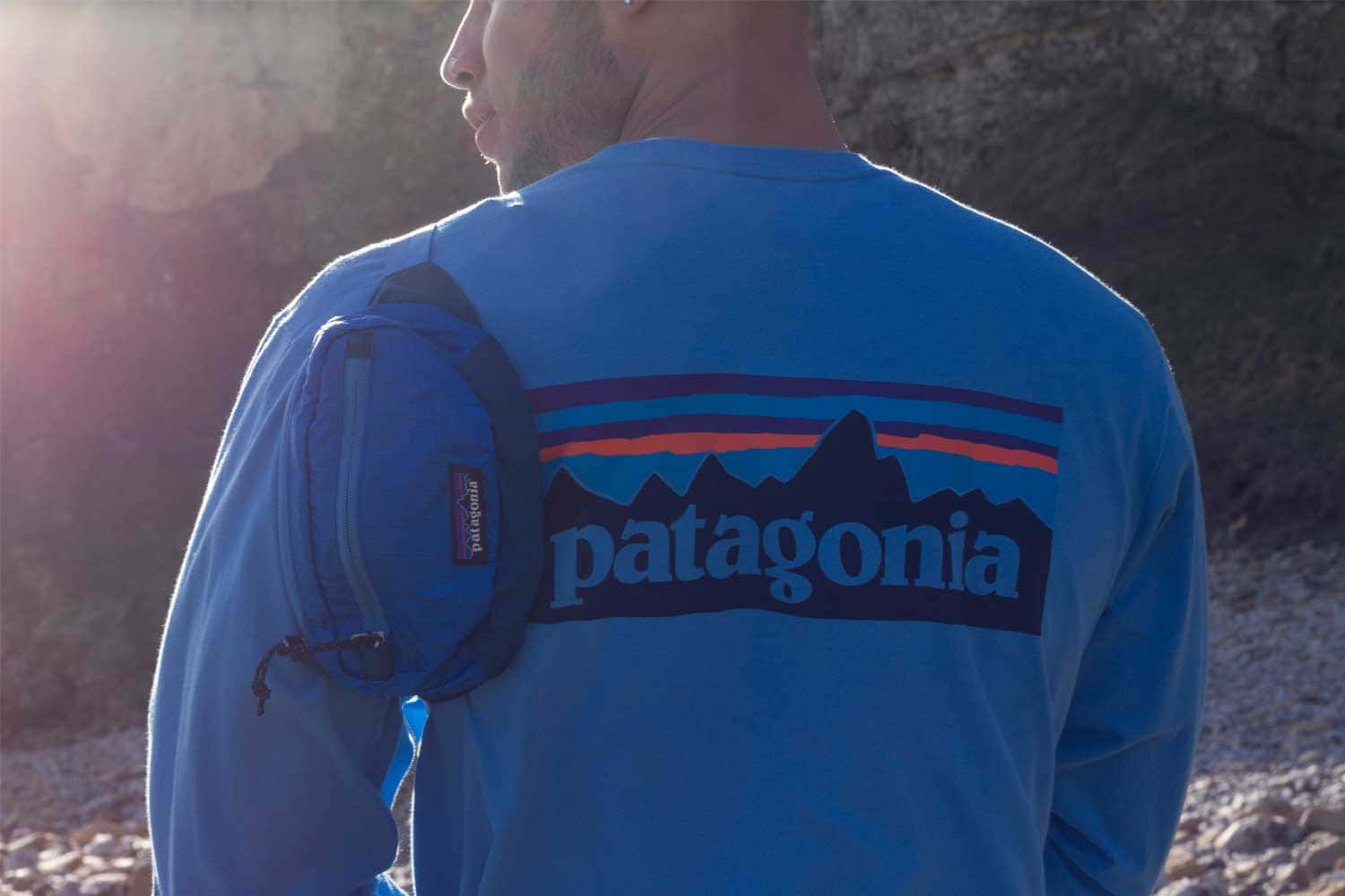 Patagonia 桌布 壁紙 — 一名身穿藍色Patagonia襯衫的男子，腰間掛著腰包，站在陽光普照的岩石背景前。