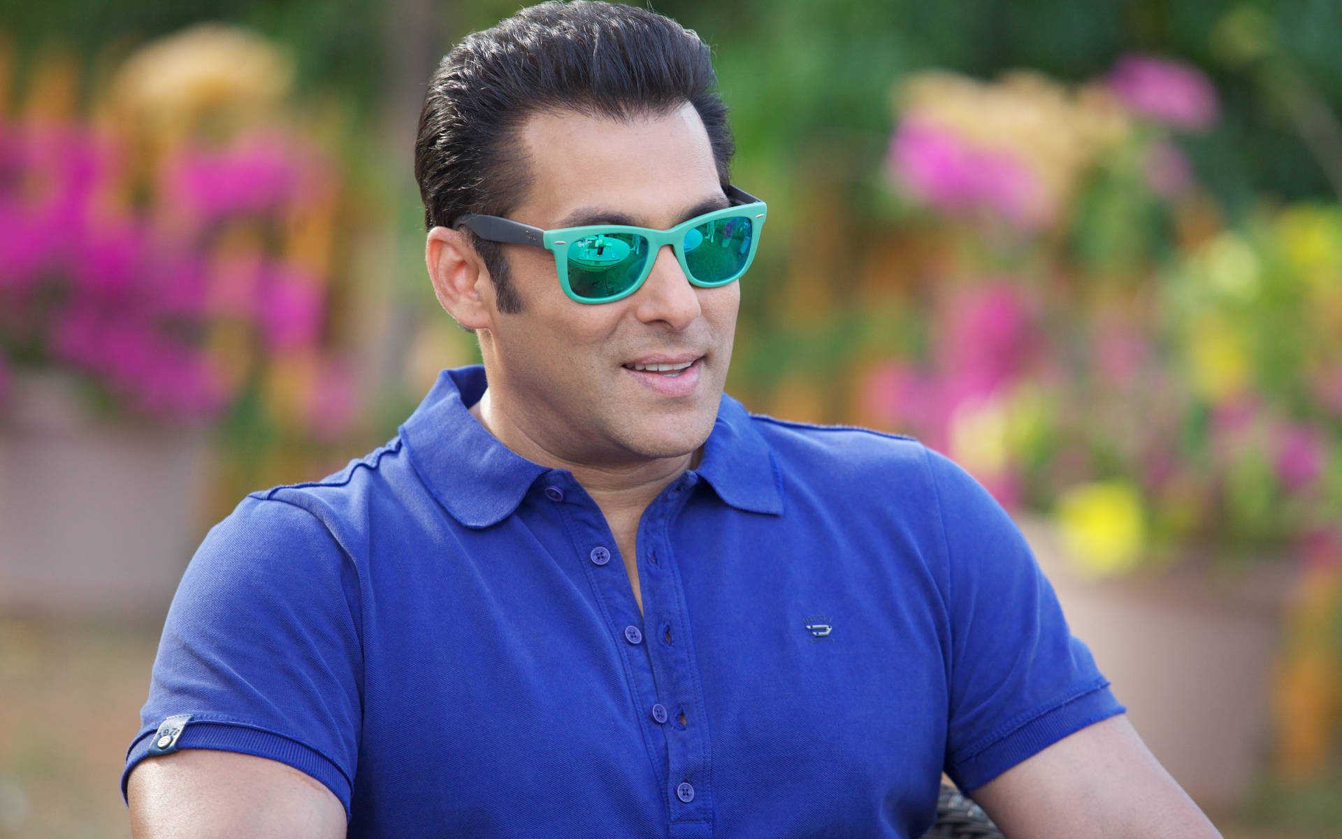 Salman Khan 桌布 壁紙 — 一名身穿藍色Polo衫和青綠色太陽眼鏡的男子，在鮮花背景前自信地微笑。