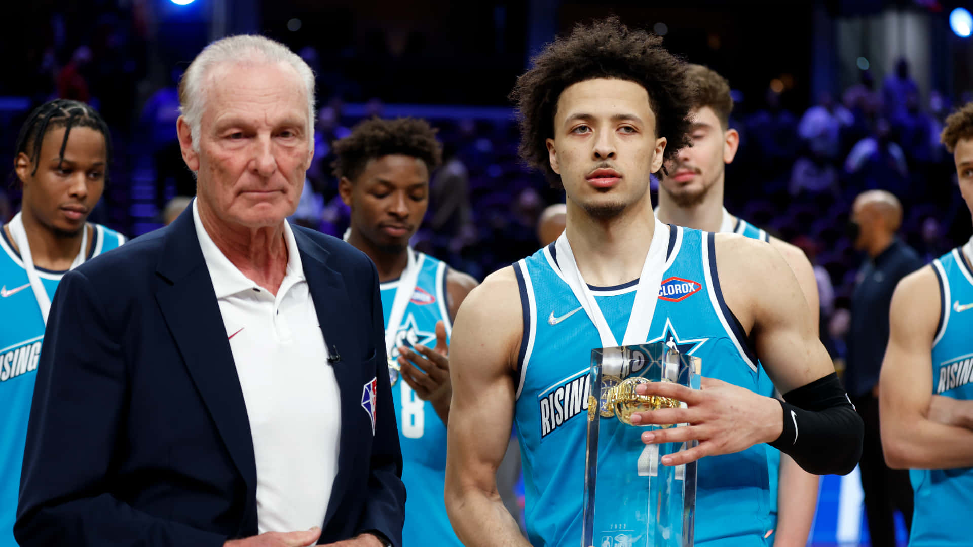 NBA 桌布 壁紙 — 一名身穿藍色Rising Stars球衣的年輕籃球運動員，在燈光明亮的場館中，手持獎盃，站在一位身穿西裝的年長男子旁邊。