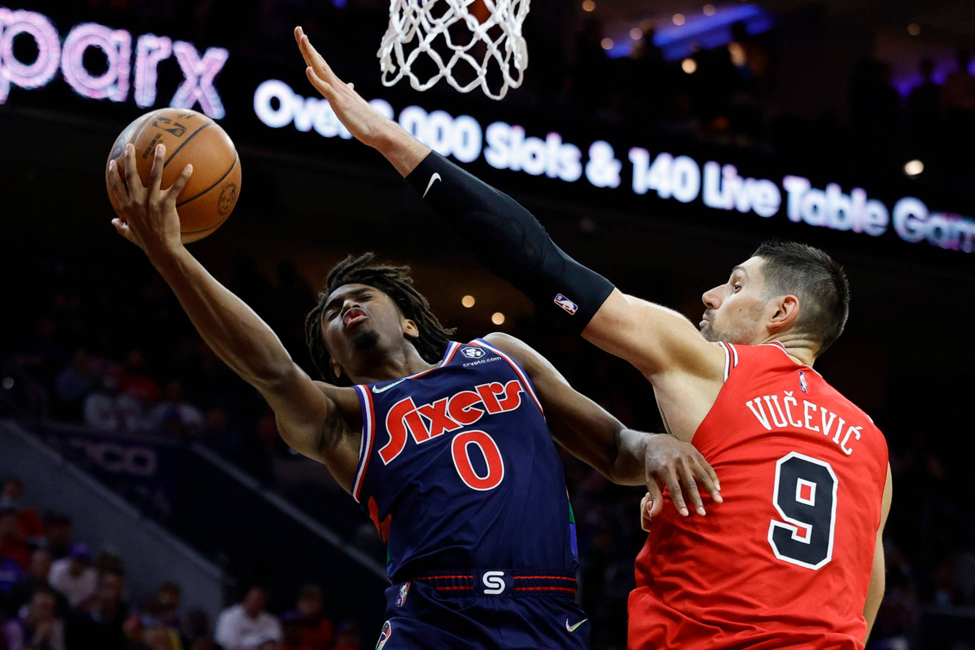 76ers 桌布 壁紙 — 一名身穿藍色Sixers球衣的籃球運動員跳起投籃，而一名紅色球衣的防守球員在明亮的場館燈光下阻擋他的路線。