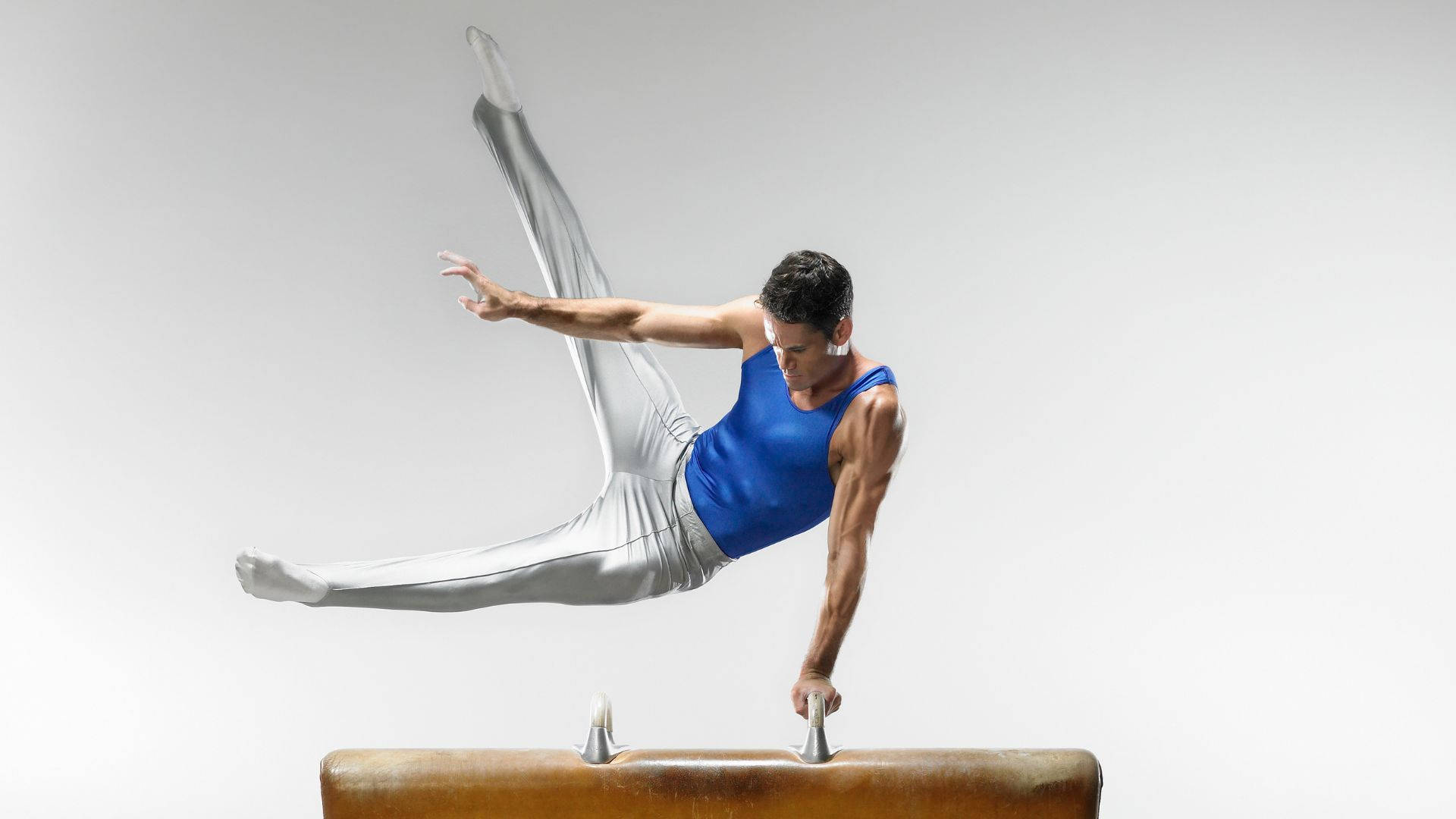 Athletic 桌布 壁紙 — 一名身穿藍色上衣和銀色褲子的男性體操運動員在白色背景的木製鞍馬上做出動態姿勢。