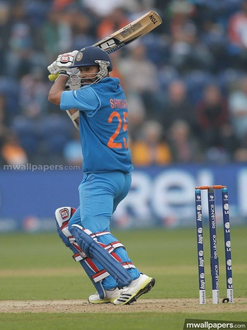 shikhar dhawan 桌布 壁紙 — 一名身穿藍色制服的板球擊球手在綠色場地上的比賽中強力揮棒擊球。