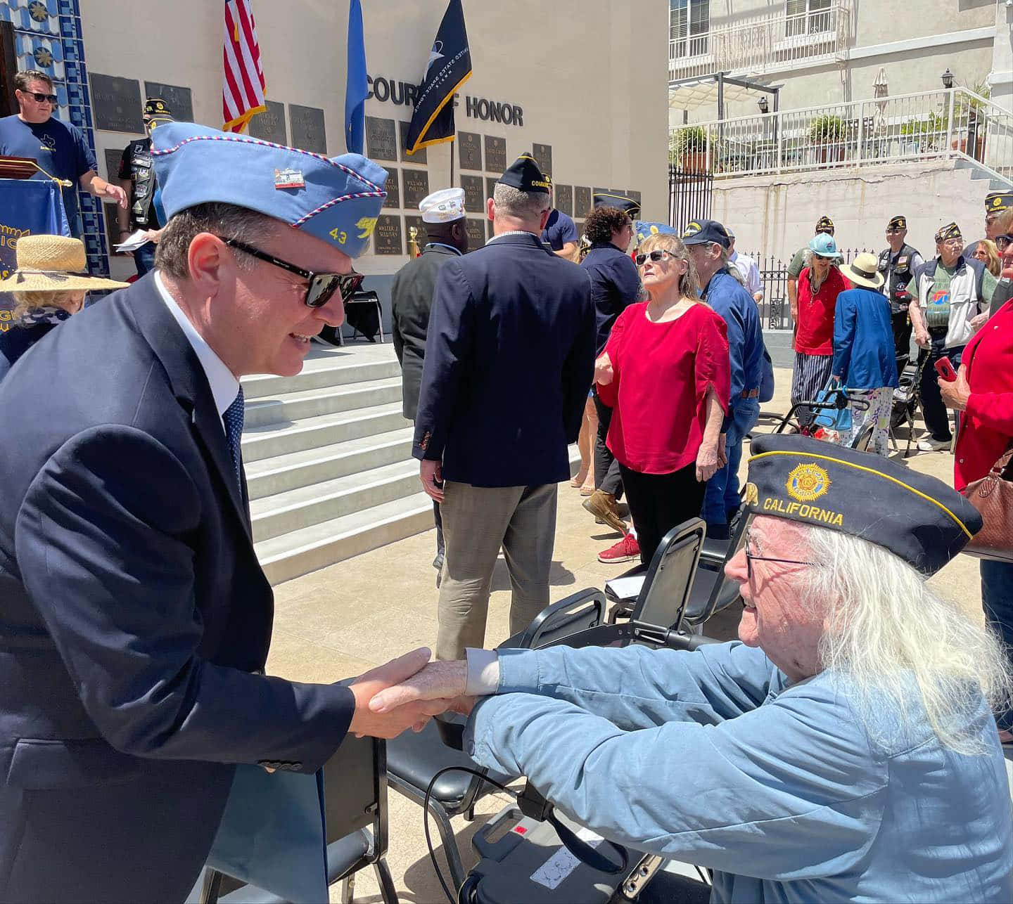Veterans Day 桌布 壁紙 — 一名身穿藍色制服的男士在陽光明媚的戶外儀式中與坐輪椅的退伍軍人握手。