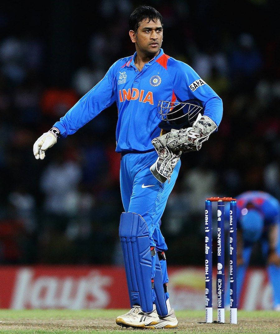 MS Dhoni 桌布 壁紙 — 一名身穿藍色印度隊服的板球運動員在夜間燈光球場上，手持頭盔和手套站在三柱門附近。