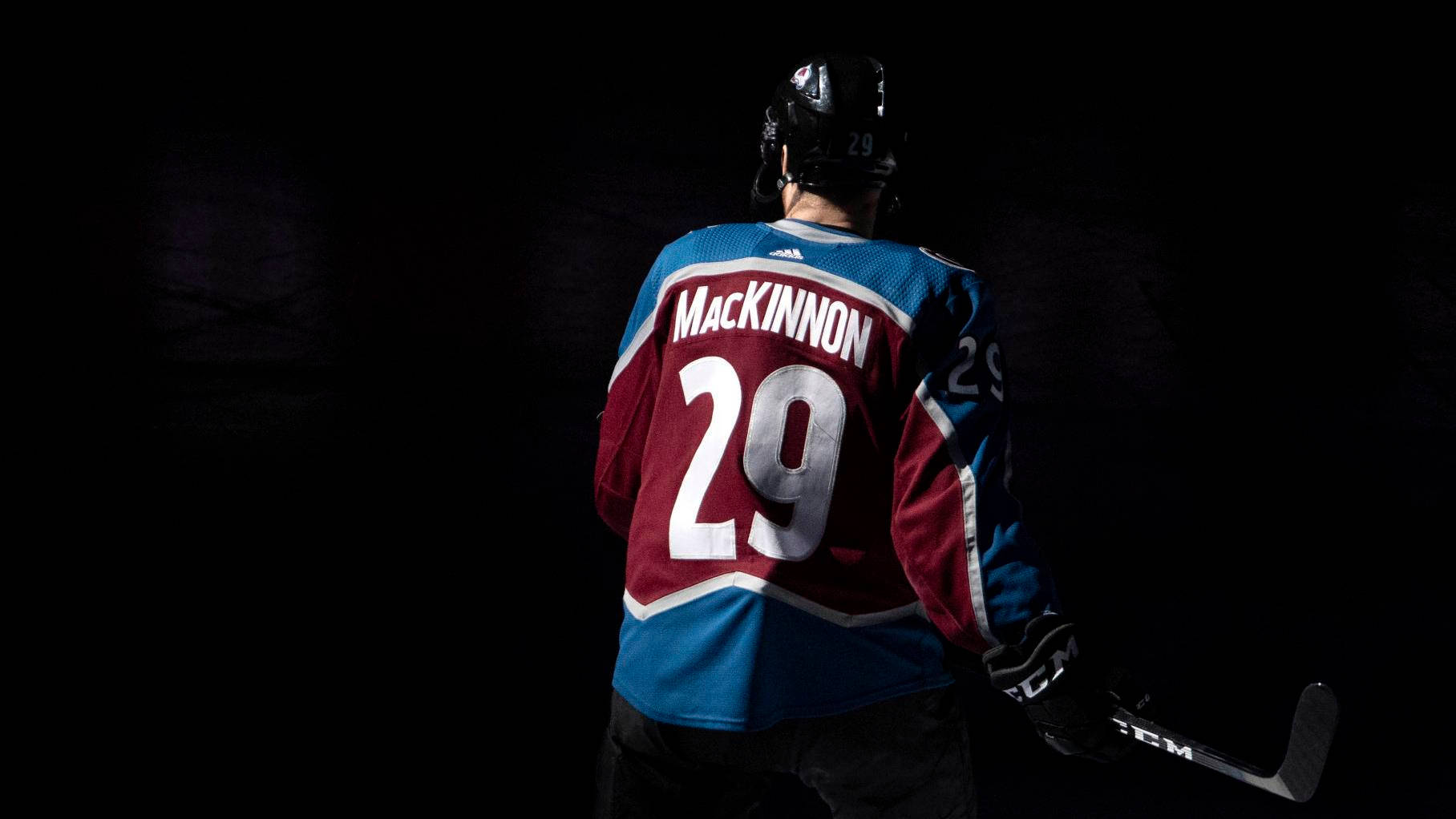 運動 桌布 壁紙 — 一名身穿藍色和栗色球衣、名字為MacKinnon、號碼29的冰球運動員站在黑暗中，手持球棍。