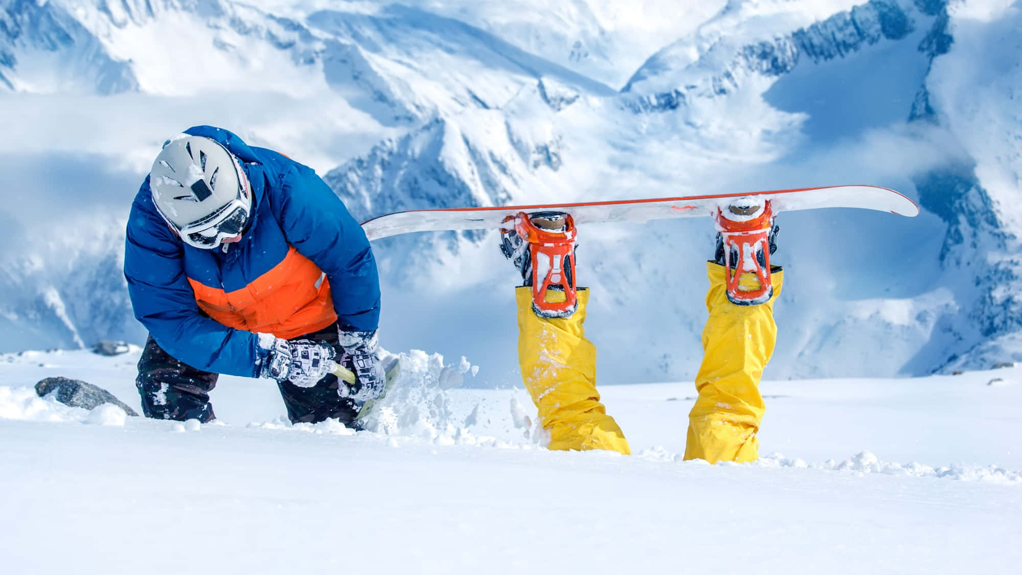 Snowboarding 桌布 壁紙 — 一名身穿藍色和橙色裝備的滑雪板運動員彎腰，而另一名穿著黃色褲子的人在新鮮的粉雪中直立拿著滑雪板，背景是白雪覆蓋的山脈。