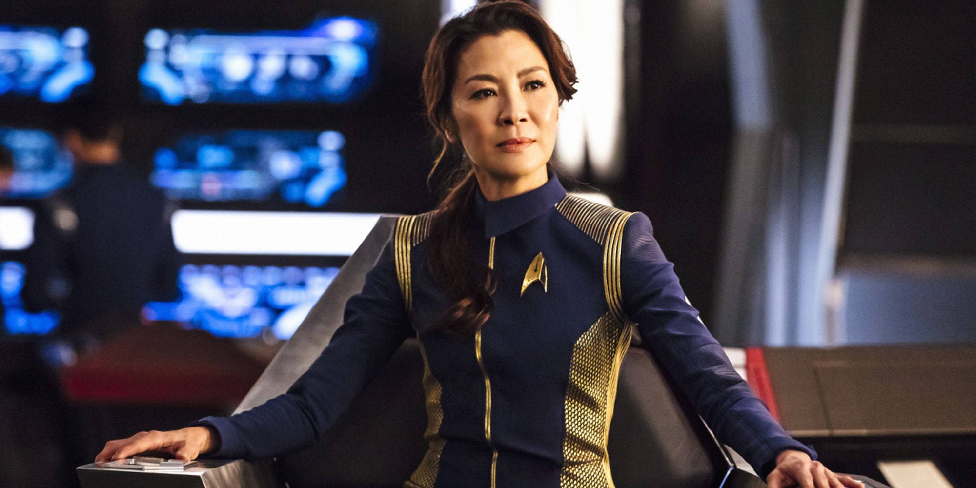 Discovery 桌布 壁紙 — 一名身穿藍色和金色制服的星際艦隊軍官，平靜地坐在一張未來風格的指揮椅上，背後有發光的藍色螢幕。