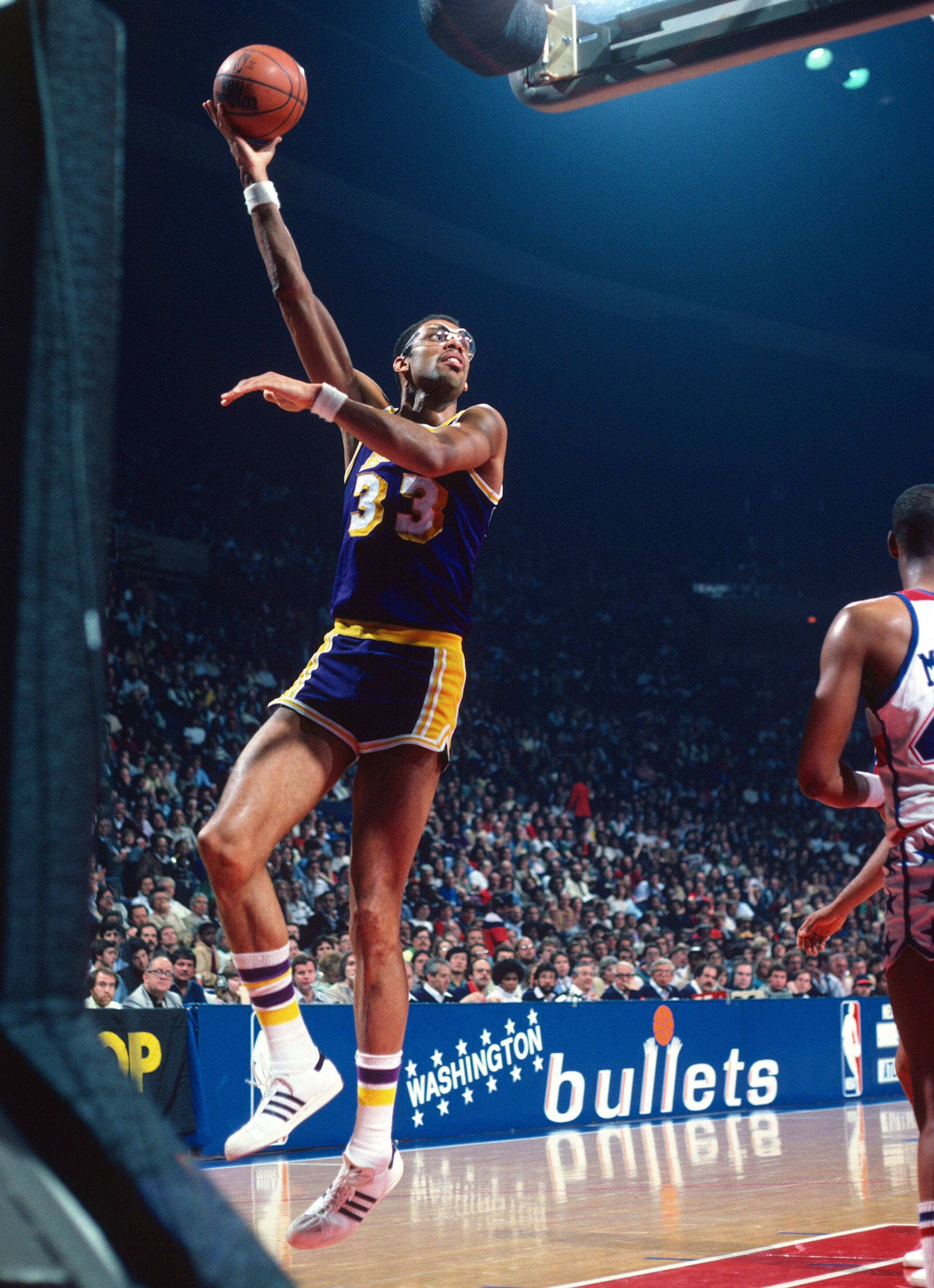 Kareem 桌布 壁紙 — 一名身穿藍色和金色球衣的籃球運動員在明亮的場館燈光下比賽中高高躍起投籃。