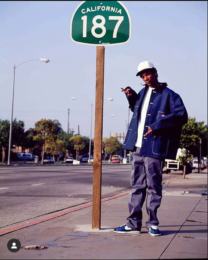 Young Snoop Dogg 桌布 壁紙 — 一名身穿藍色外套、戴白色帽子的年輕男子，自信地站在陽光明媚的城市人行道上，旁邊是加州187高速公路標誌。