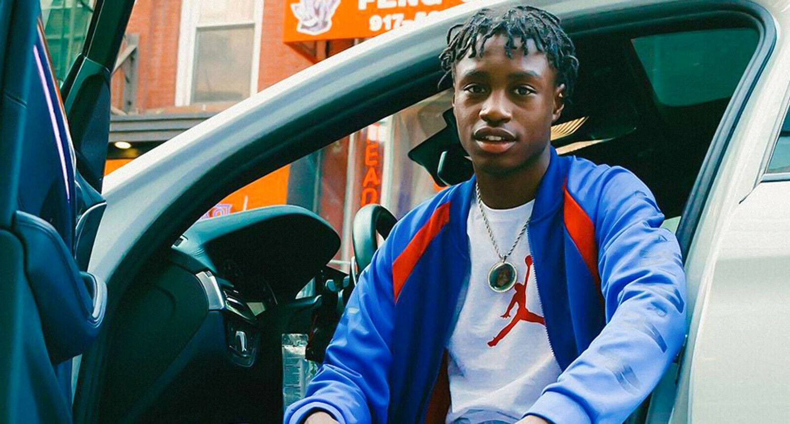 Lil Tjay 桌布 壁紙 — 一名身穿藍色外套和Jordan T恤的年輕男子坐在車內，車門敞開，背景是城市風光。