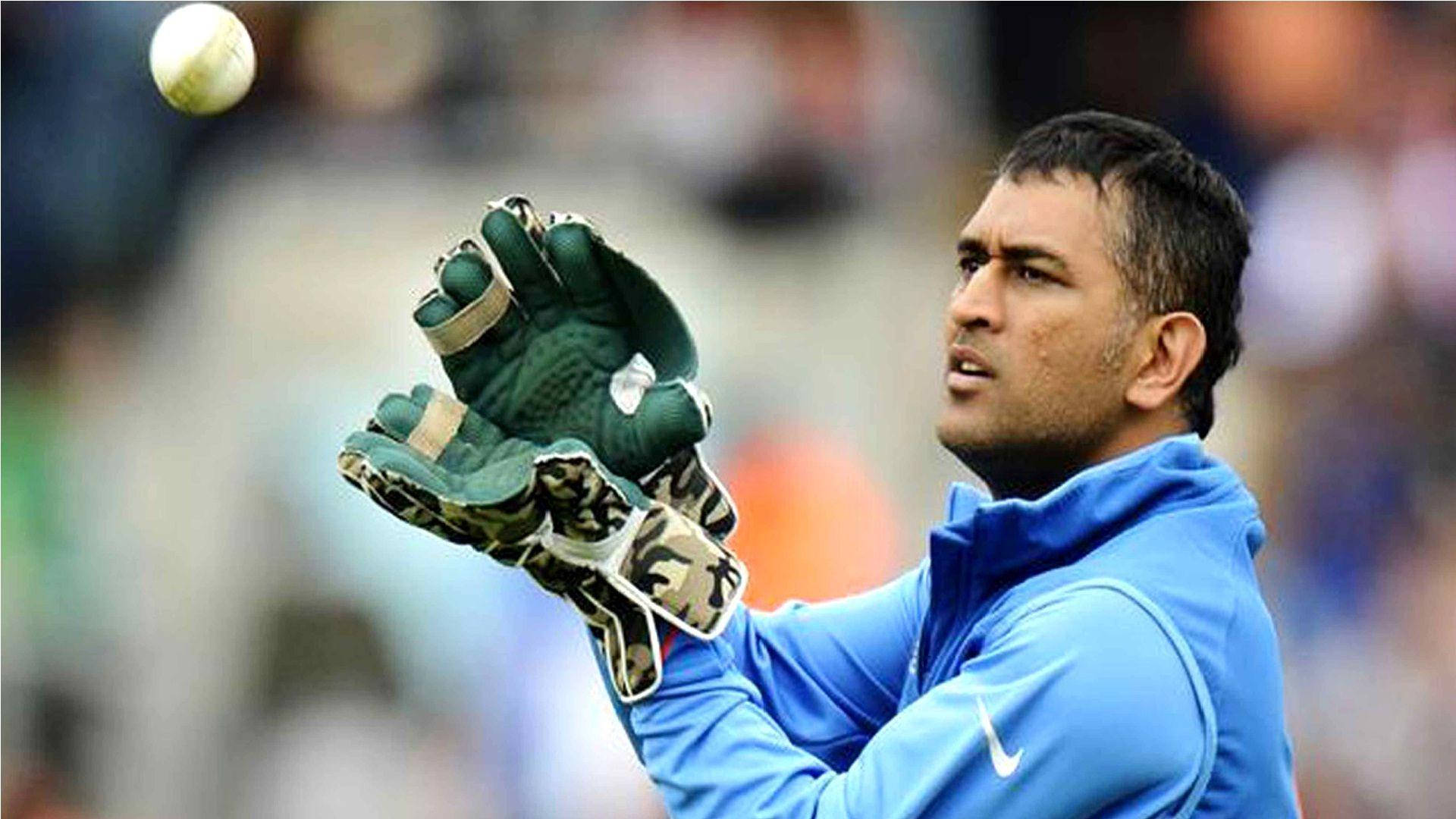 Ms Dhoni 桌布 壁紙 — 一名身穿藍色外套和綠色手套的板球選手，全神貫注地伸手接住空中的白色板球。
