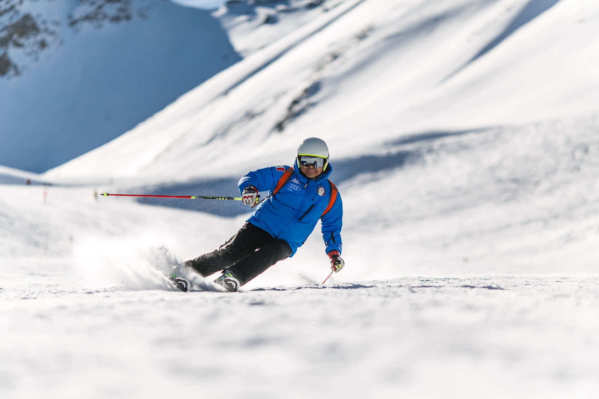 ski 桌布 壁紙 — 一名身穿藍色外套的滑雪者，在明亮的陽光下滑下白雪覆蓋的山坡，揚起粉雪。