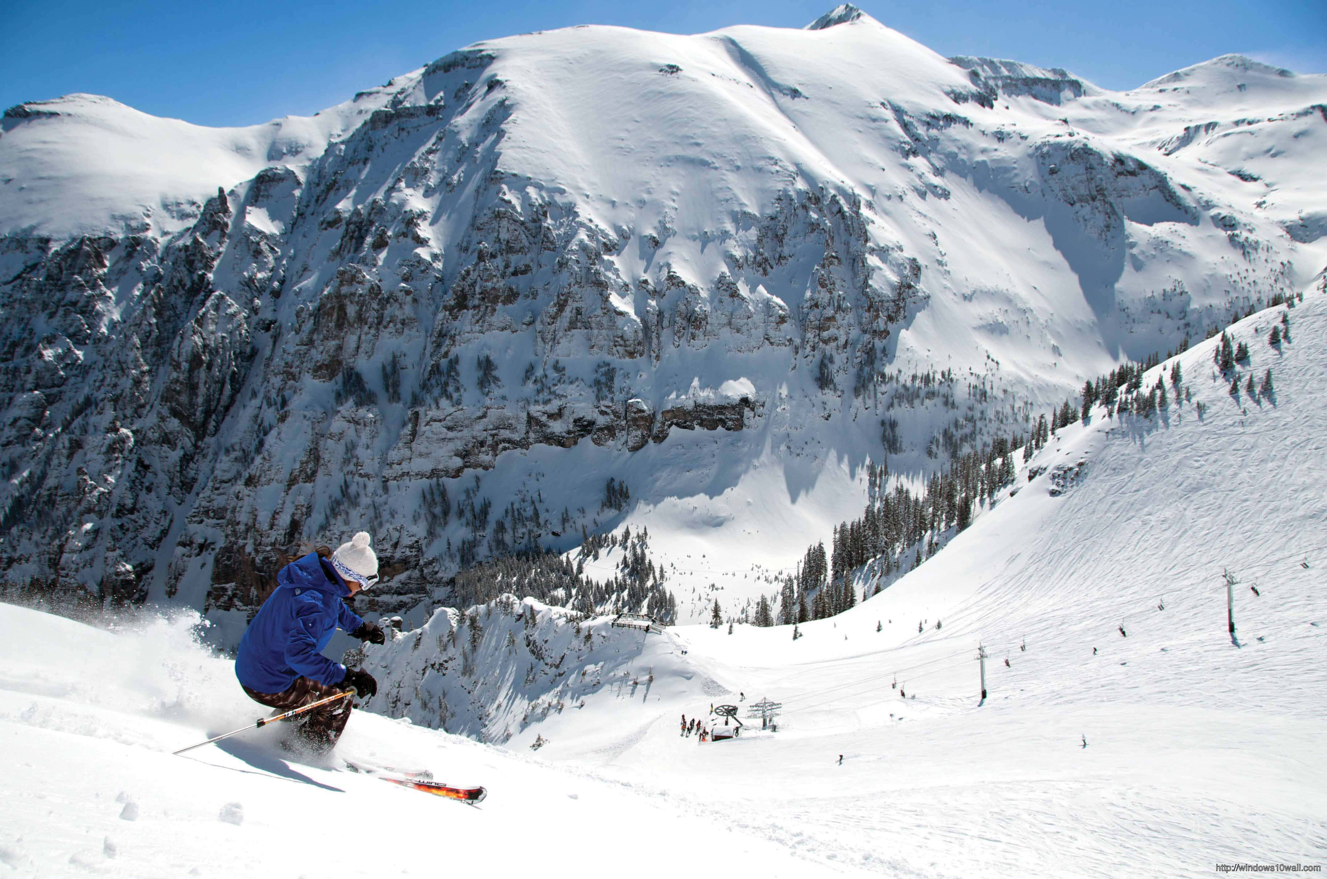 Aspen Colorado 桌布 壁紙 — 一名身穿藍色外套的滑雪者，在晴朗藍天下，沿著積雪的山坡滑下，周圍有崎嶇的山峰和常青樹。