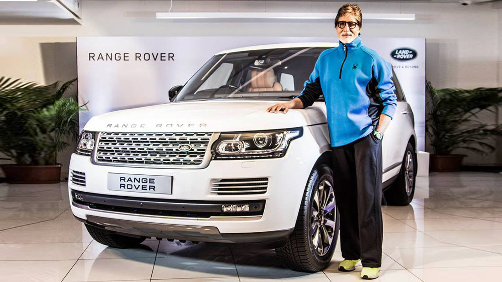 Amitabh Bachchan 桌布 壁紙 — 一名身穿藍色外套的男子自信地站在一輛白色Range Rover旁邊，身處明亮的展示間，室內有盆栽植物和品牌牆壁。
