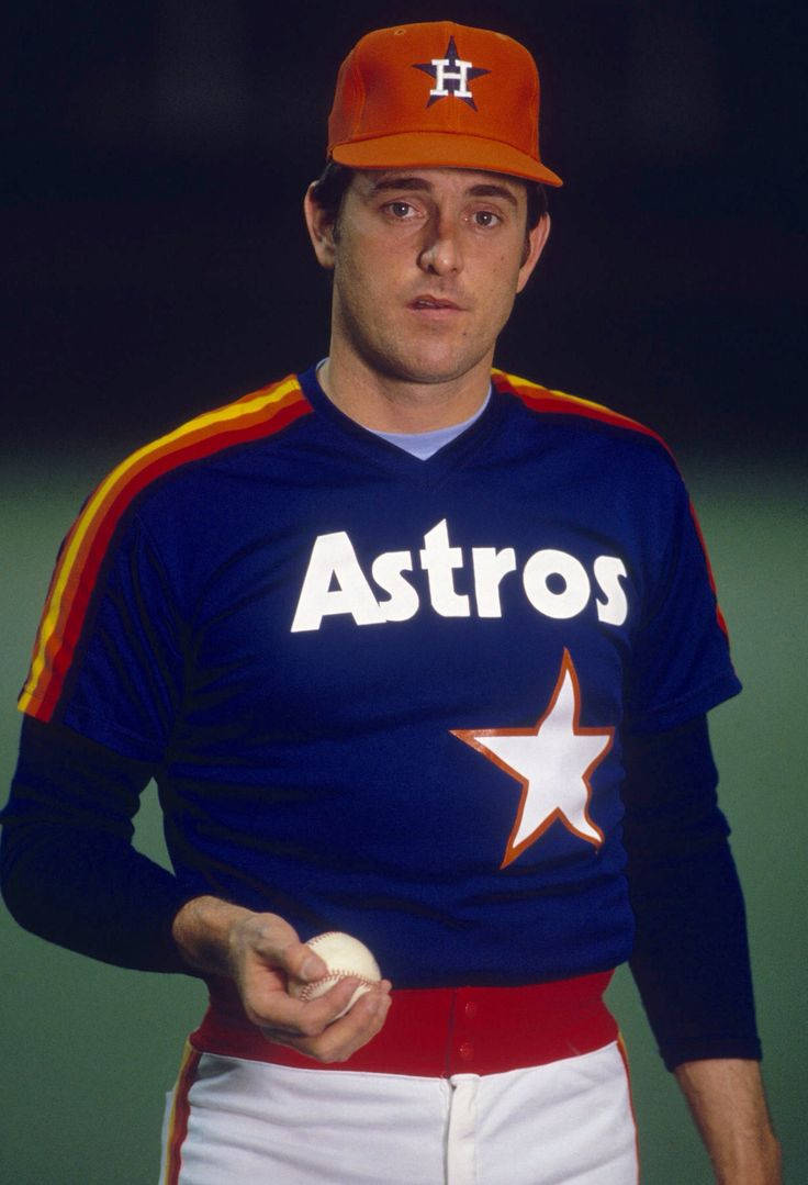 Nolan Ryan 桌布 壁紙 — 一名身穿藍色太空人球衣（帶橙色星星）並戴帽子的棒球選手，在深綠色背景前手持棒球。