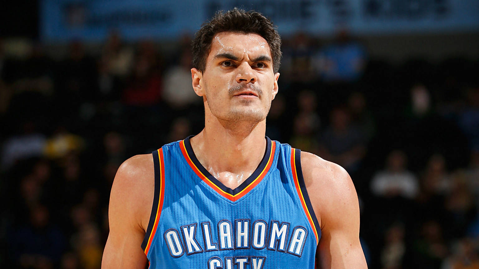 Steven Adams 桌布 壁紙 — 一名身穿藍色奧克拉荷馬城球衣的專注籃球運動員，在明亮的場館燈光下於比賽中凝視著。