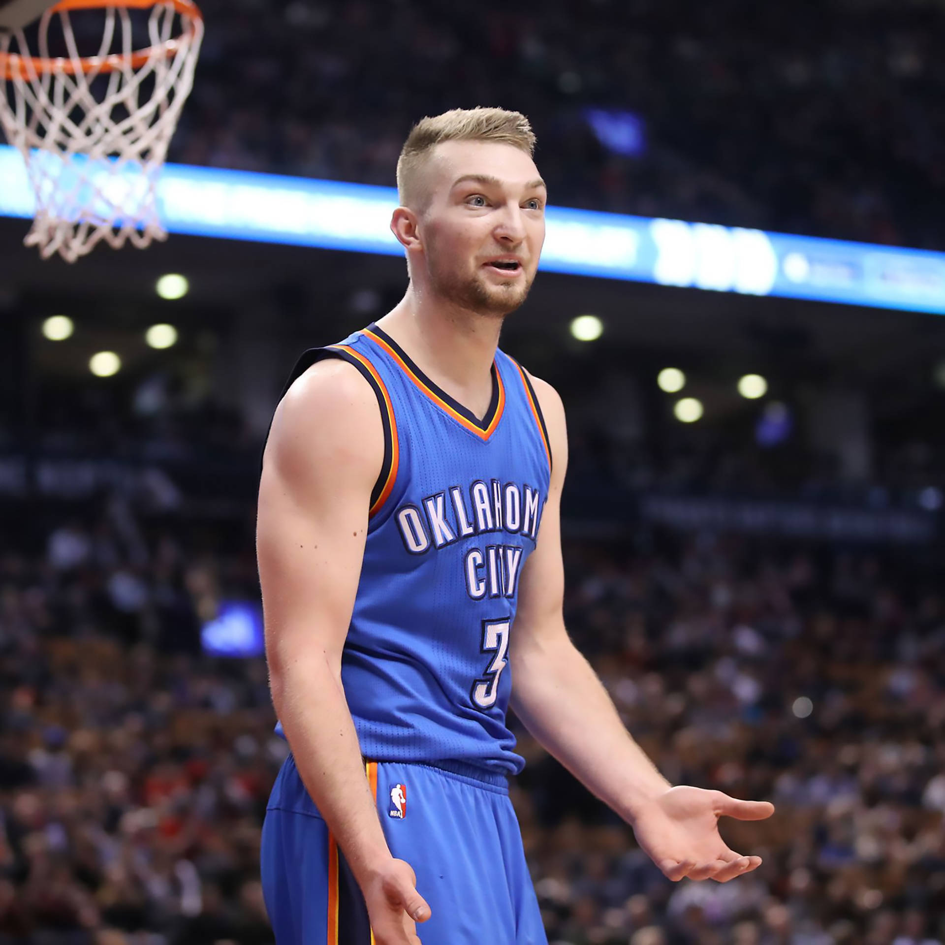 domantas sabonis 桌布 壁紙 — 一名身穿藍色奧克拉荷馬城球衣的籃球運動員在擠滿觀眾的場館中，張開雙手站在籃框下。