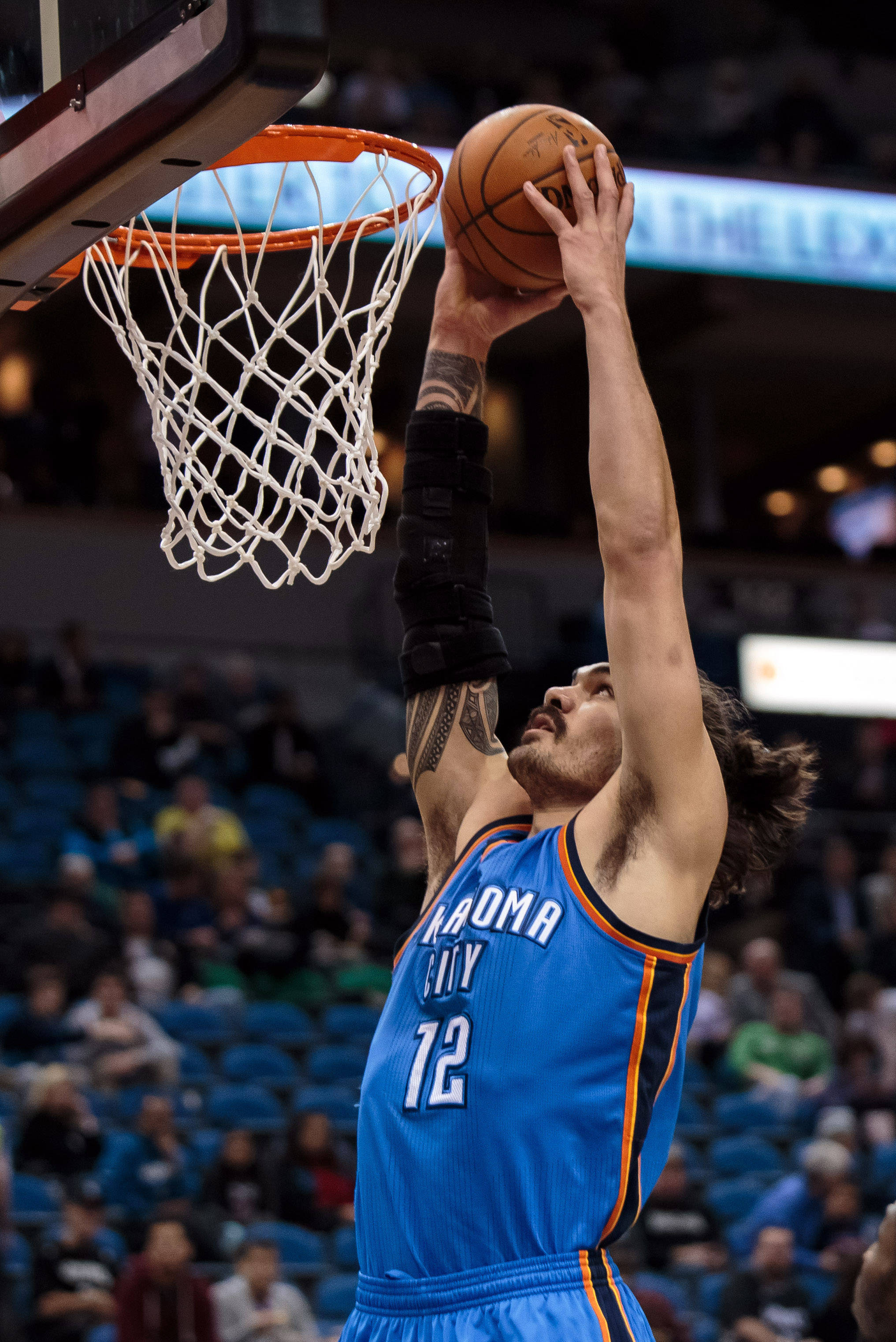 Steven Adams 桌布 壁紙 — 一名身穿藍色奧克拉荷馬城球衣的籃球運動員在比賽中灌籃，手臂上的刺青清晰可見。