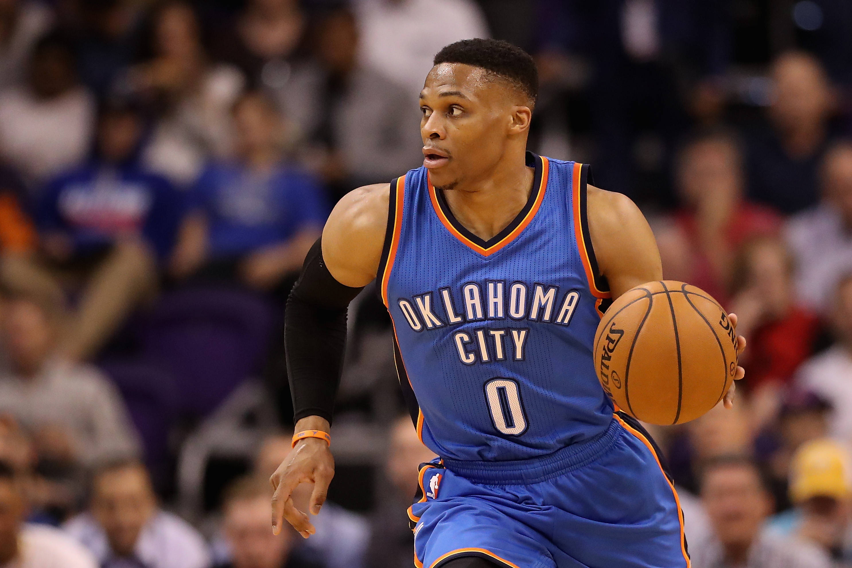 Westbrook 桌布 壁紙 — 一名身穿藍色奧克拉荷馬城球衣的籃球運動員在比賽中激烈運球，身後是模糊的人群。