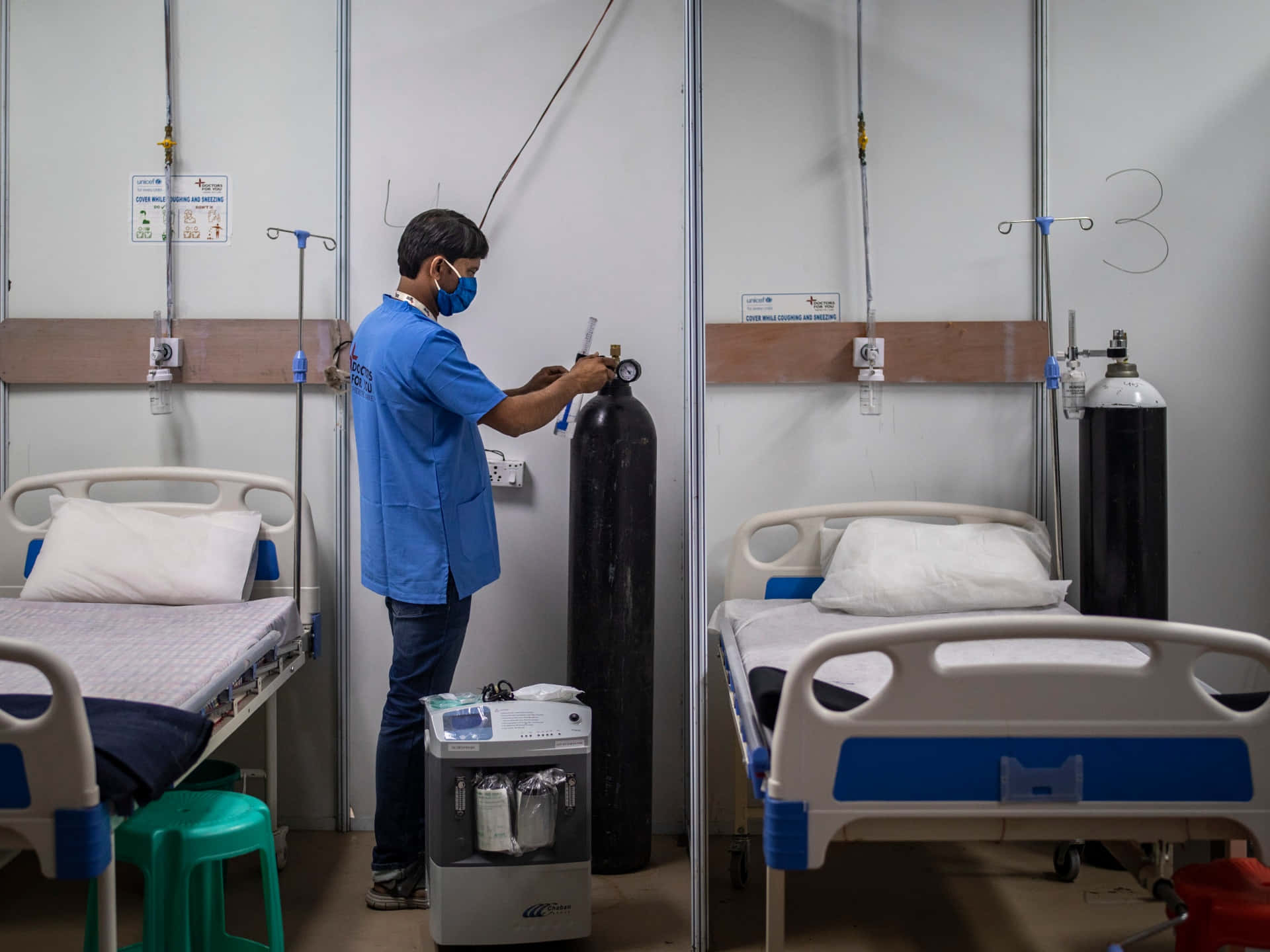 Hospital 桌布 壁紙 — 一名身穿藍色手術衣及口罩的醫護人員，在臨時病房的兩張病床之間調整氧氣罐。