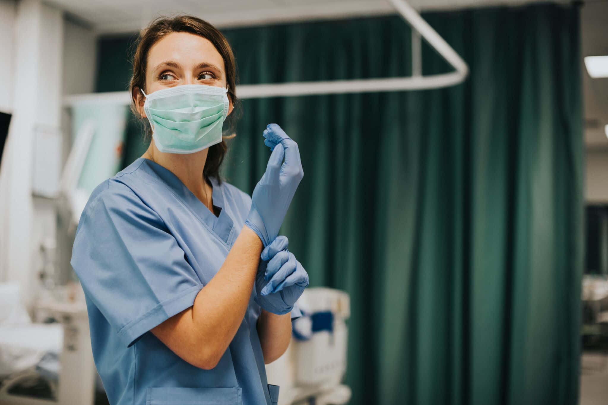Nursing 桌布 壁紙 — 一名身穿藍色手術衣和口罩的女護士在帶有綠色窗簾的醫院房間內小心地戴上藍色醫用手套。