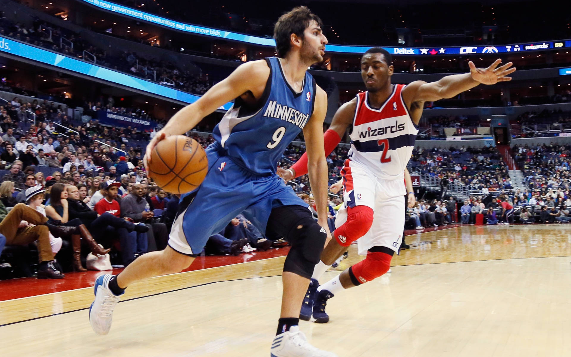 Timberwolves 桌布 壁紙 — 一名身穿藍色明尼蘇達球衣的籃球運動員，在擠滿觀眾的場館中運球越過華盛頓巫師隊的防守球員。