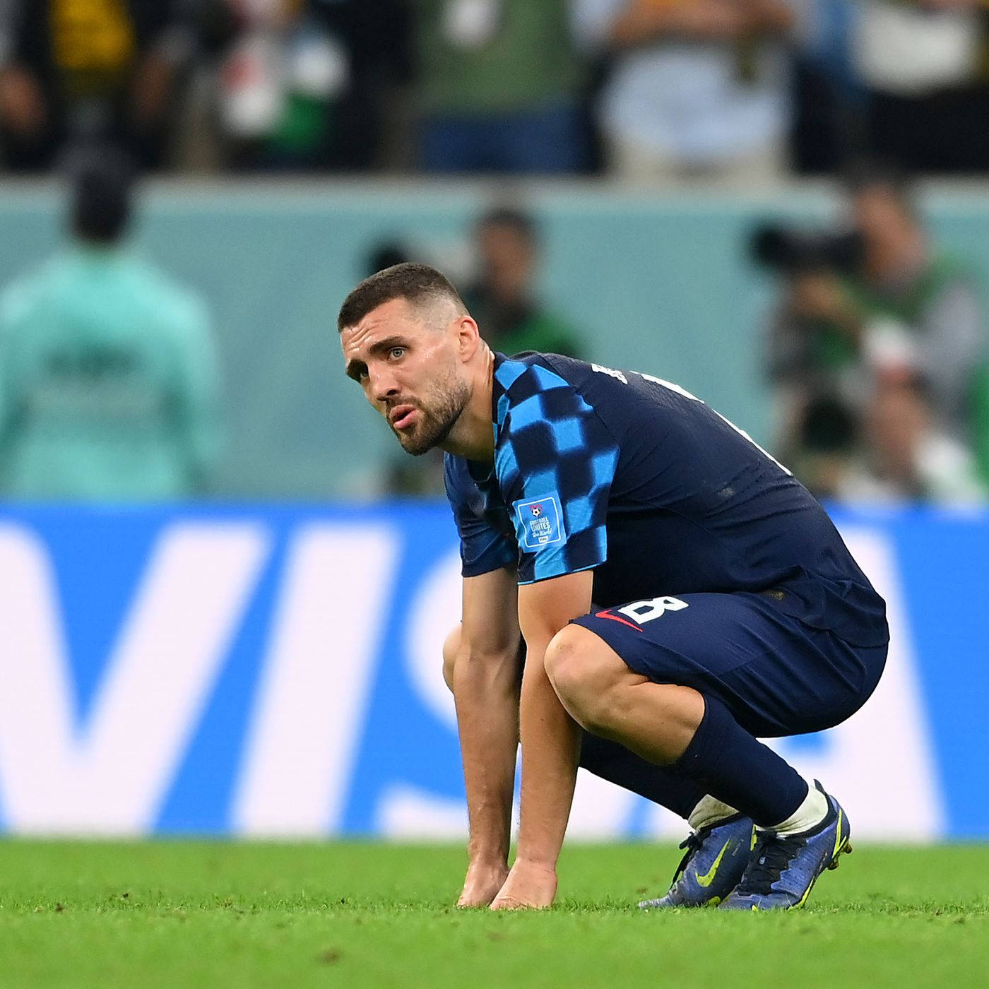 Football Players 桌布 壁紙 — 一名身穿藍色格子球衣的足球員在比賽期間，帶著專注的表情跪在綠色球場上。