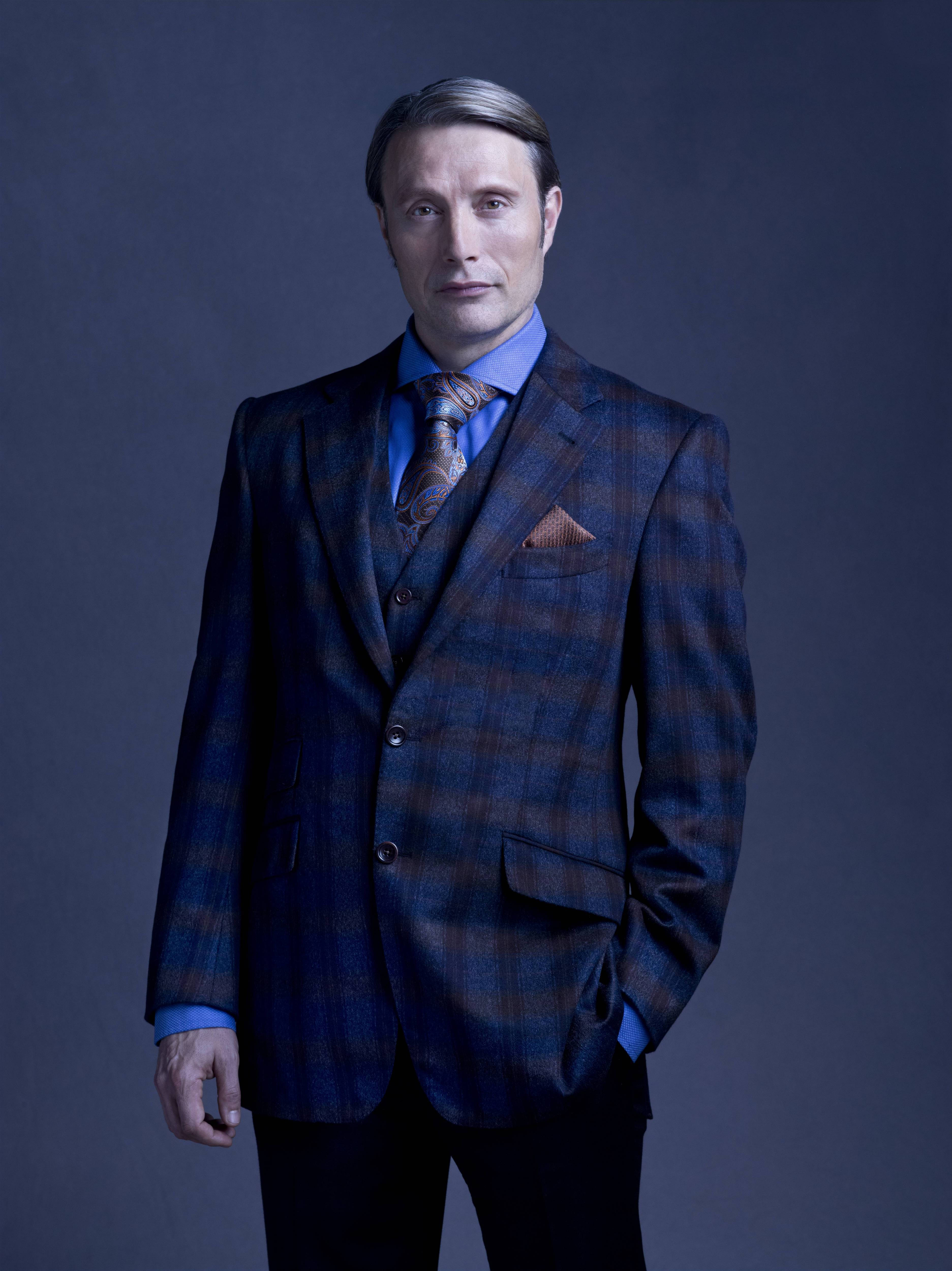 Mads Mikkelsen 桌布 壁紙 — 一名身穿藍色格紋西裝、搭配圖案領帶的男子，在深灰色攝影棚背景前自信地站立。