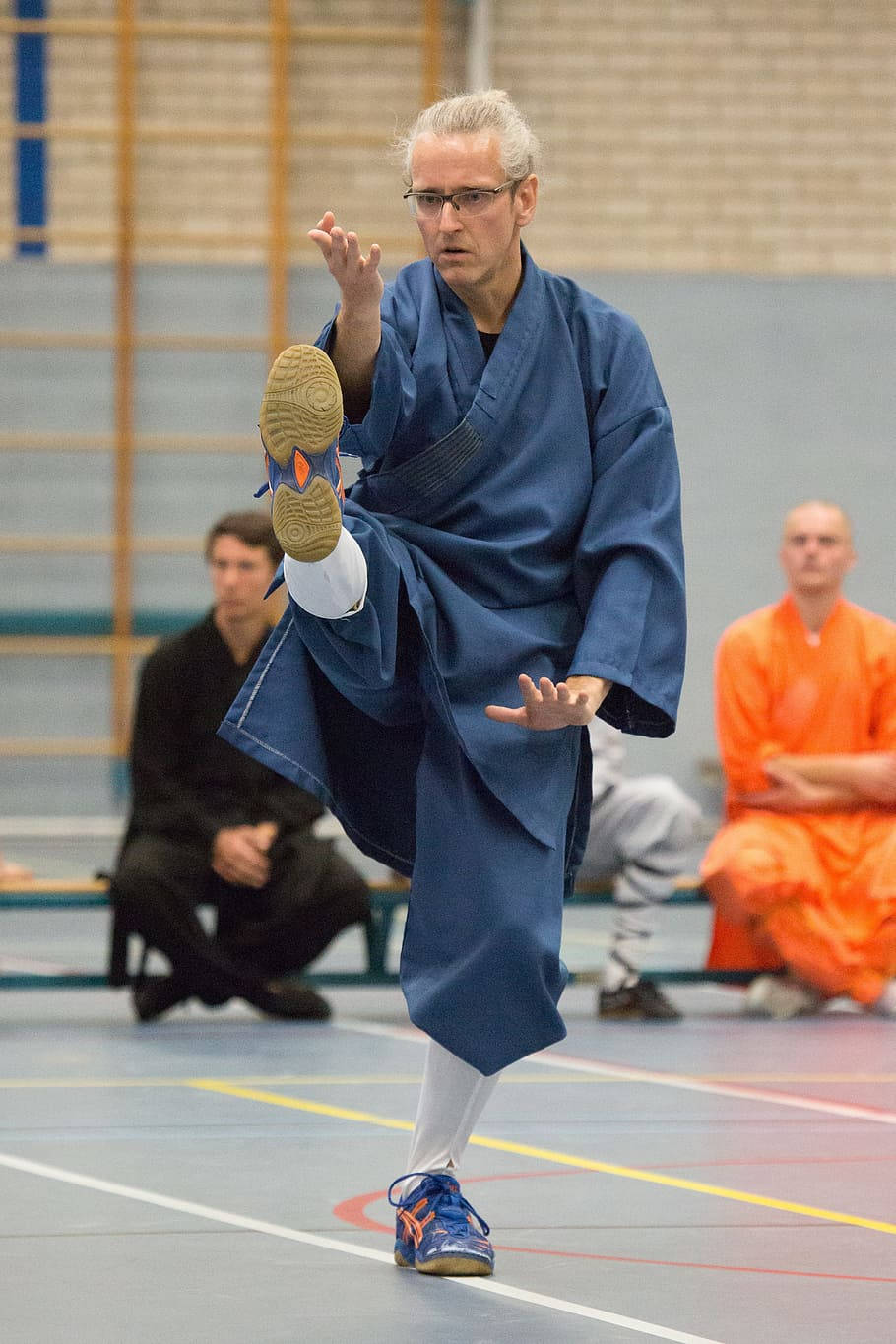 martial arts 桌布 壁紙 — 一名身穿藍色武術道服的男子在體育館內進行高踢，附近有穿著橙色和黑色長袍的觀眾坐著。