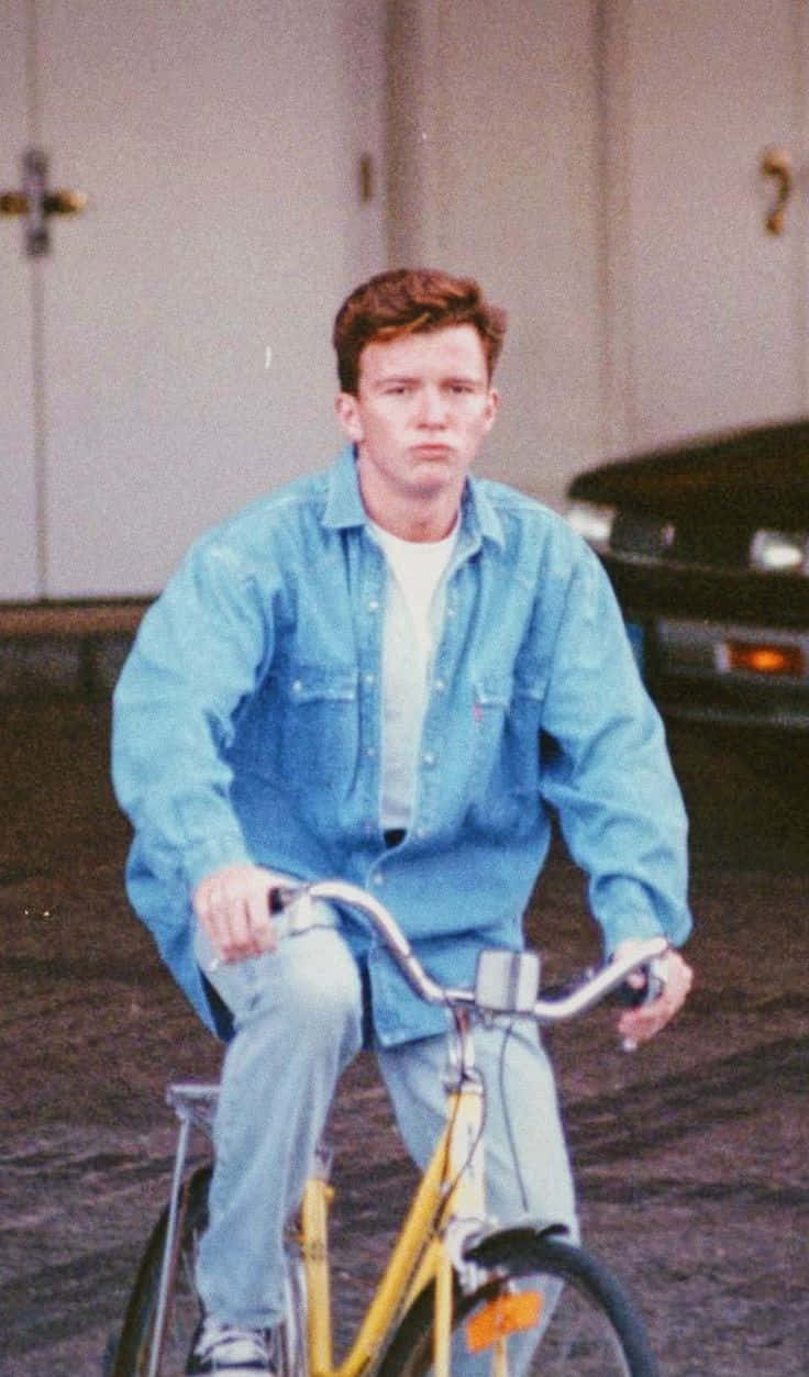 Rick Astley 桌布 壁紙 — 一名身穿藍色牛仔襯衫的年輕男子在車庫裡騎著一輛黃色腳踏車，表情嚴肅。