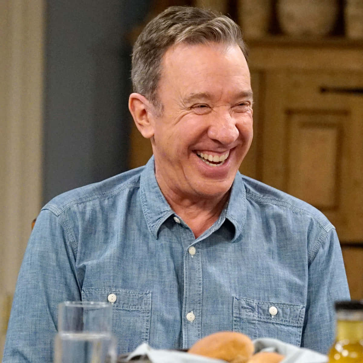 Tim Allen 桌布 壁紙 — 一名身穿藍色牛仔襯衫的男子坐在擺有食物和飲品的餐桌上開懷大笑。