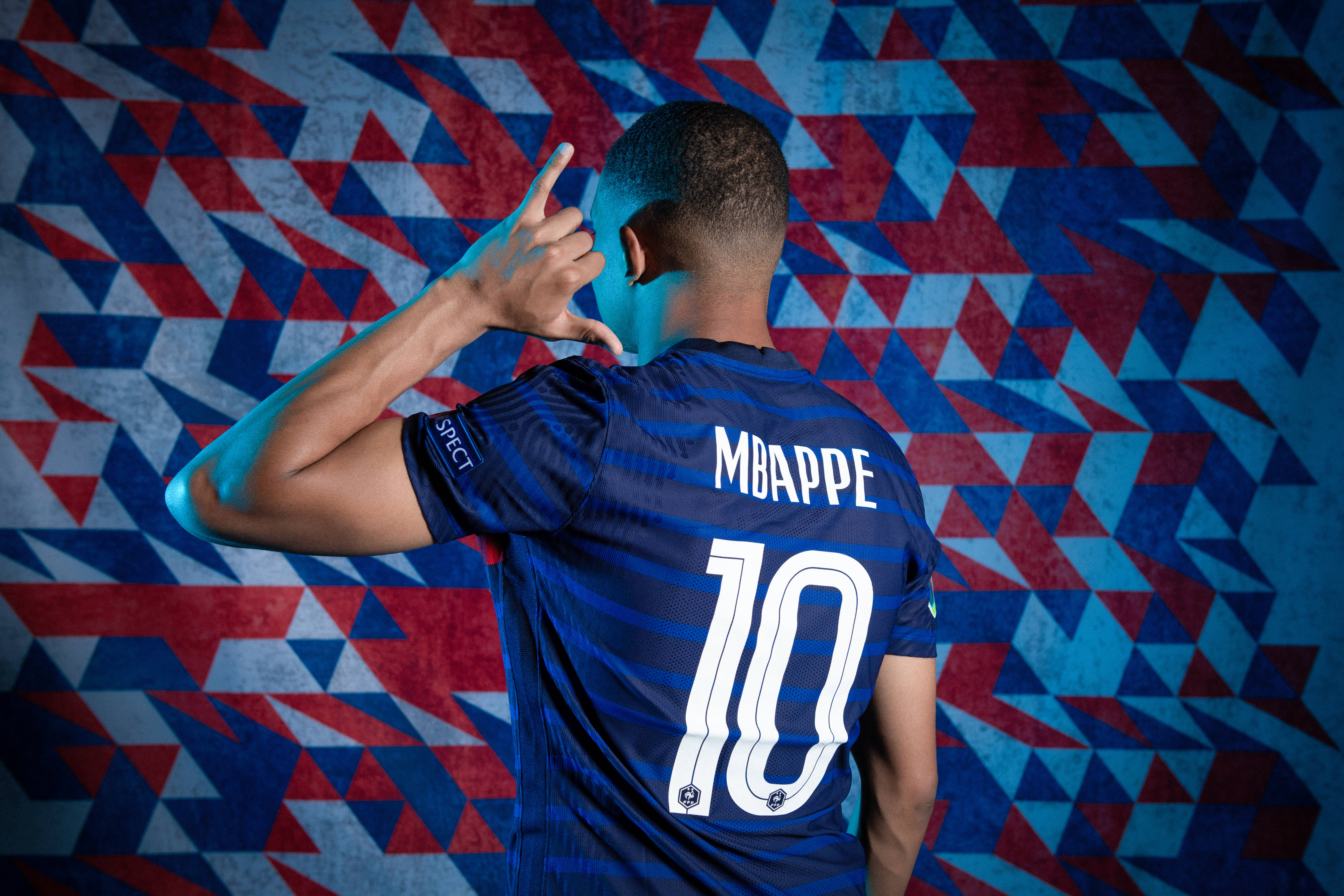 Mbappe 桌布 壁紙 — 一名身穿藍色球衣、號碼10、名字為Mbappe的足球員，在充滿活力的幾何紅、藍、白背景前做出手勢。