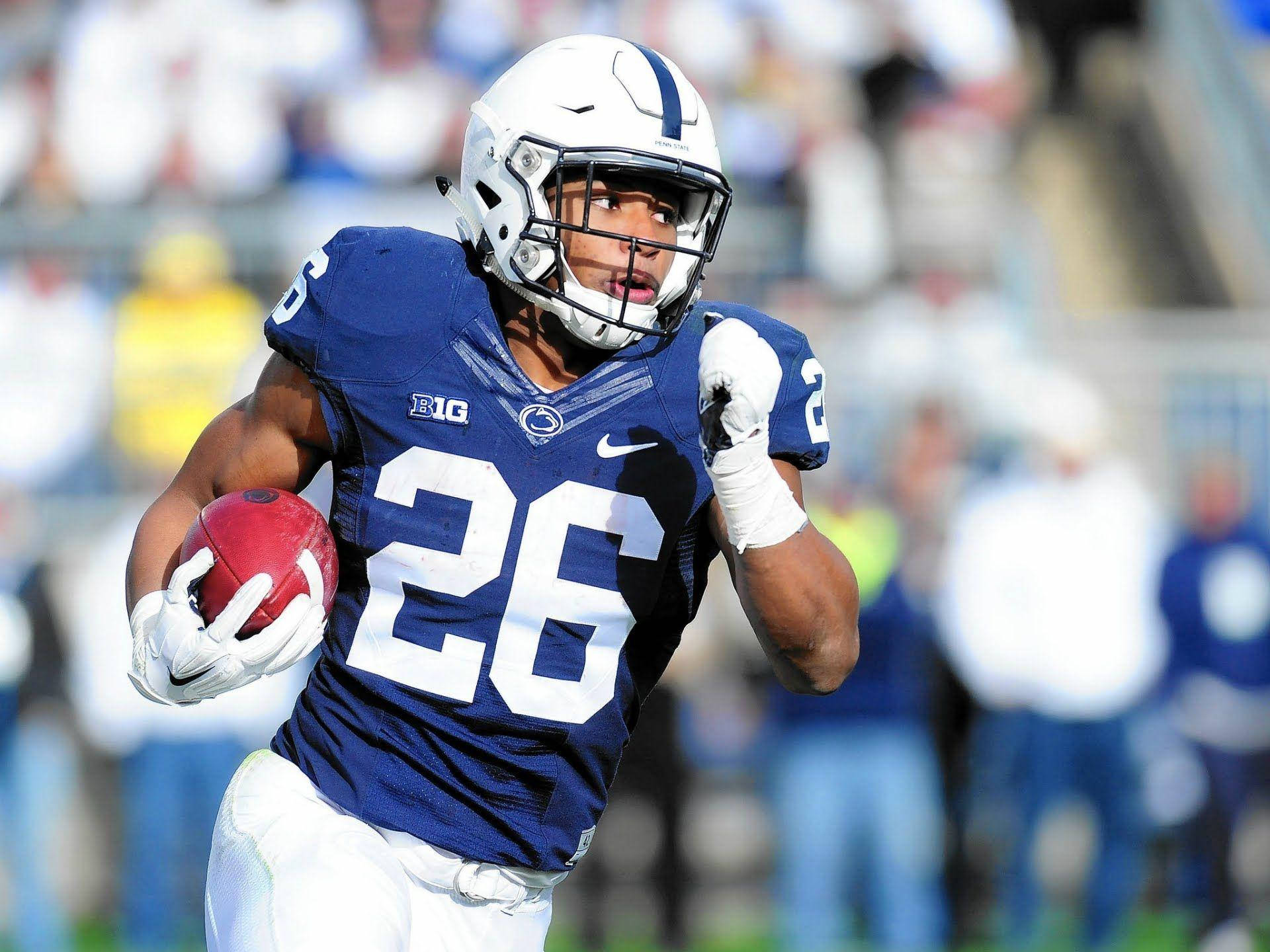Saquon Barkley 桌布 壁紙 — 一名身穿藍色球衣、號碼26的美式足球員在日間比賽中手持紅色球向前衝刺。