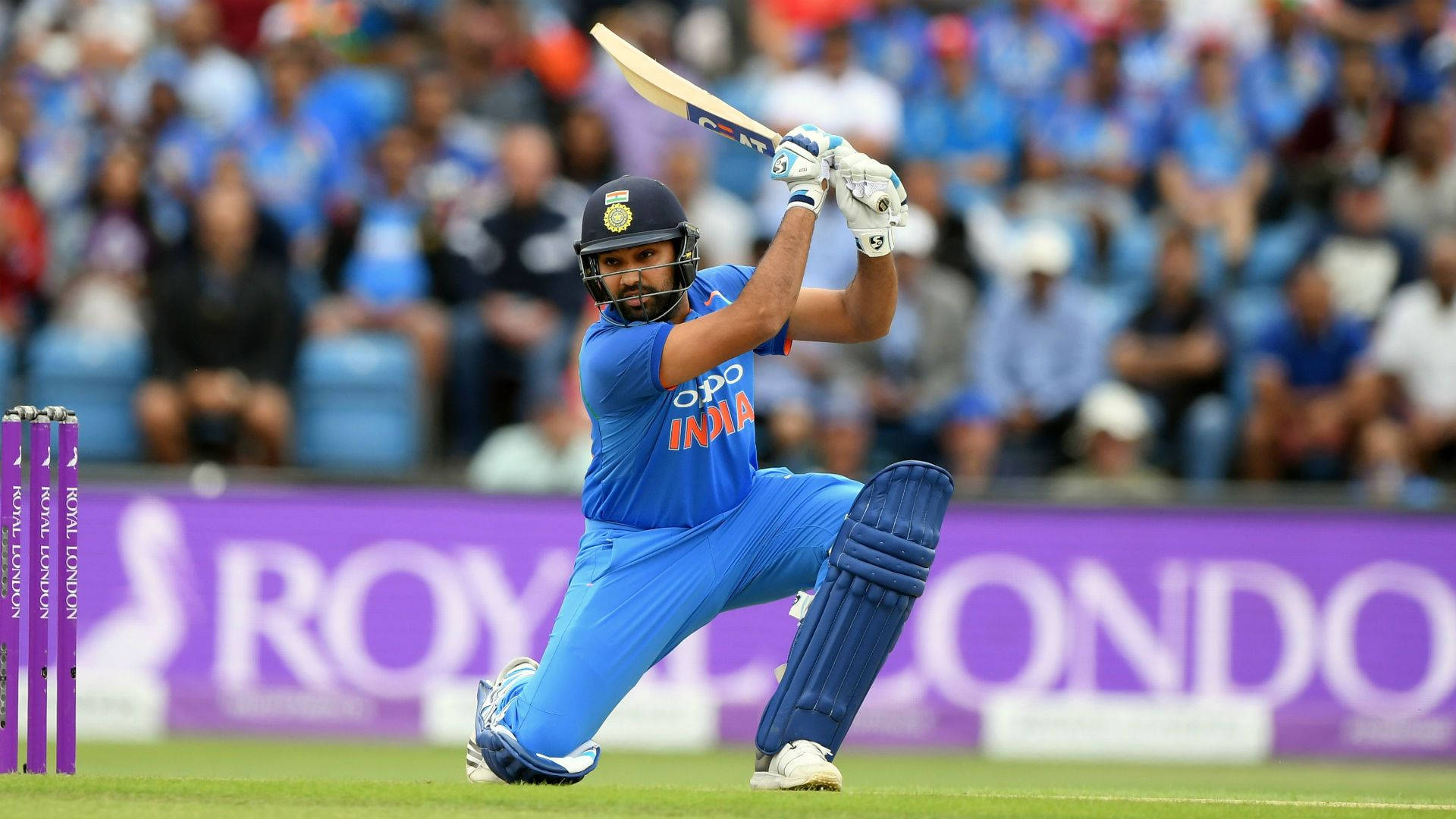 Rohit Sharma 桌布 壁紙 — 一名身穿藍色球衣的印度板球運動員在日間比賽中強力揮棒，背景有觀眾。