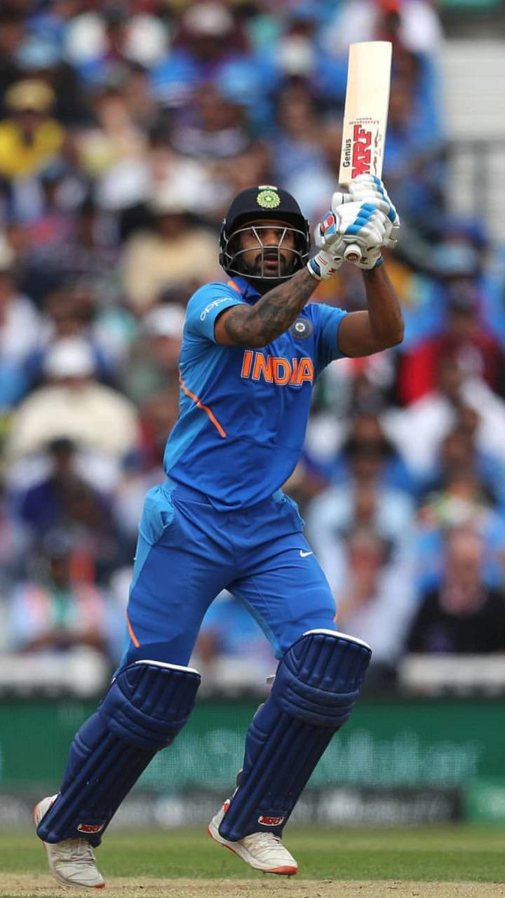 shikhar dhawan 桌布 壁紙 — 一名身穿藍色球衣的印度板球運動員在綠色球場上揮動球棒，背景是模糊的人群。