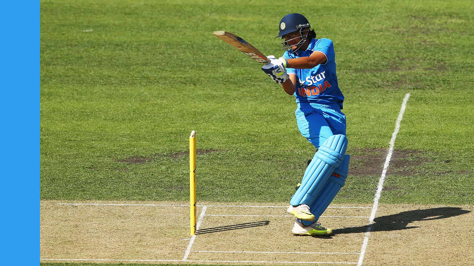 Smriti Mandhana 桌布 壁紙 — 一名身穿藍色球衣的女板球運動員在陽光明媚的綠色球場上，在三柱門附近強力揮棒。