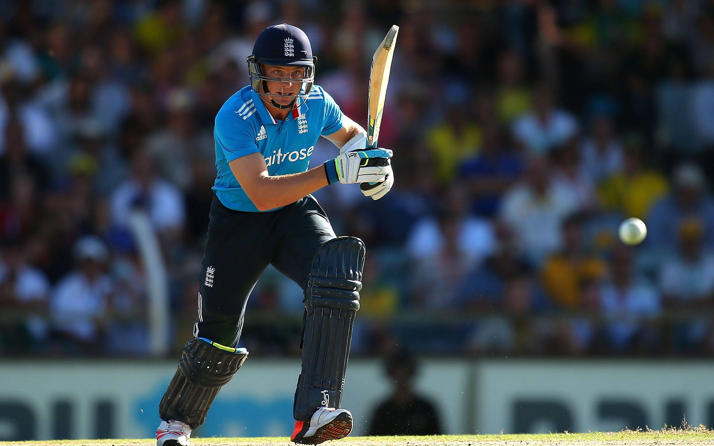 England Cricket 桌布 壁紙 — 一名身穿藍色球衣的板球擊球手在陽光明媚的比賽中揮動球棒擊打白色球，背景是模糊的觀眾。