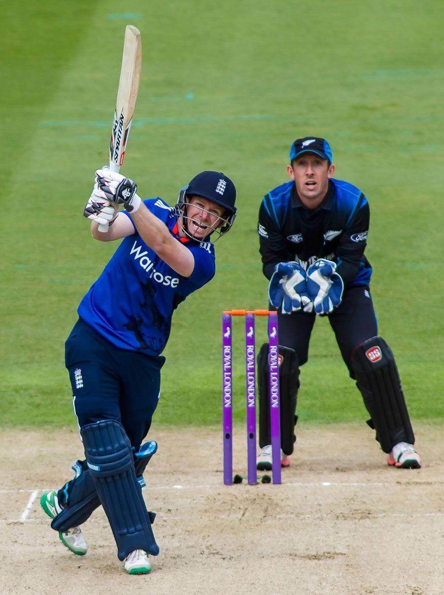 England Cricket 桌布 壁紙 — 一名身穿藍色球衣的板球擊球手強力揮棒擊球，而一名身穿深色裝備的守門員在綠色場地上的三柱門後警覺地蹲伏。