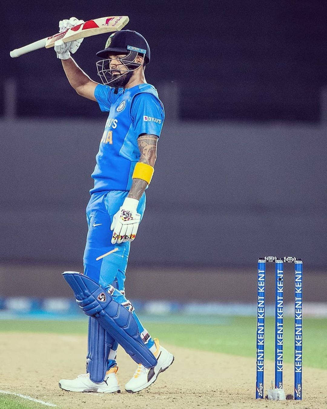 kl rahul 桌布 壁紙 — 一名身穿藍色球衣的板球運動員在球場燈光下，於三柱門旁高舉球棒慶祝勝利。